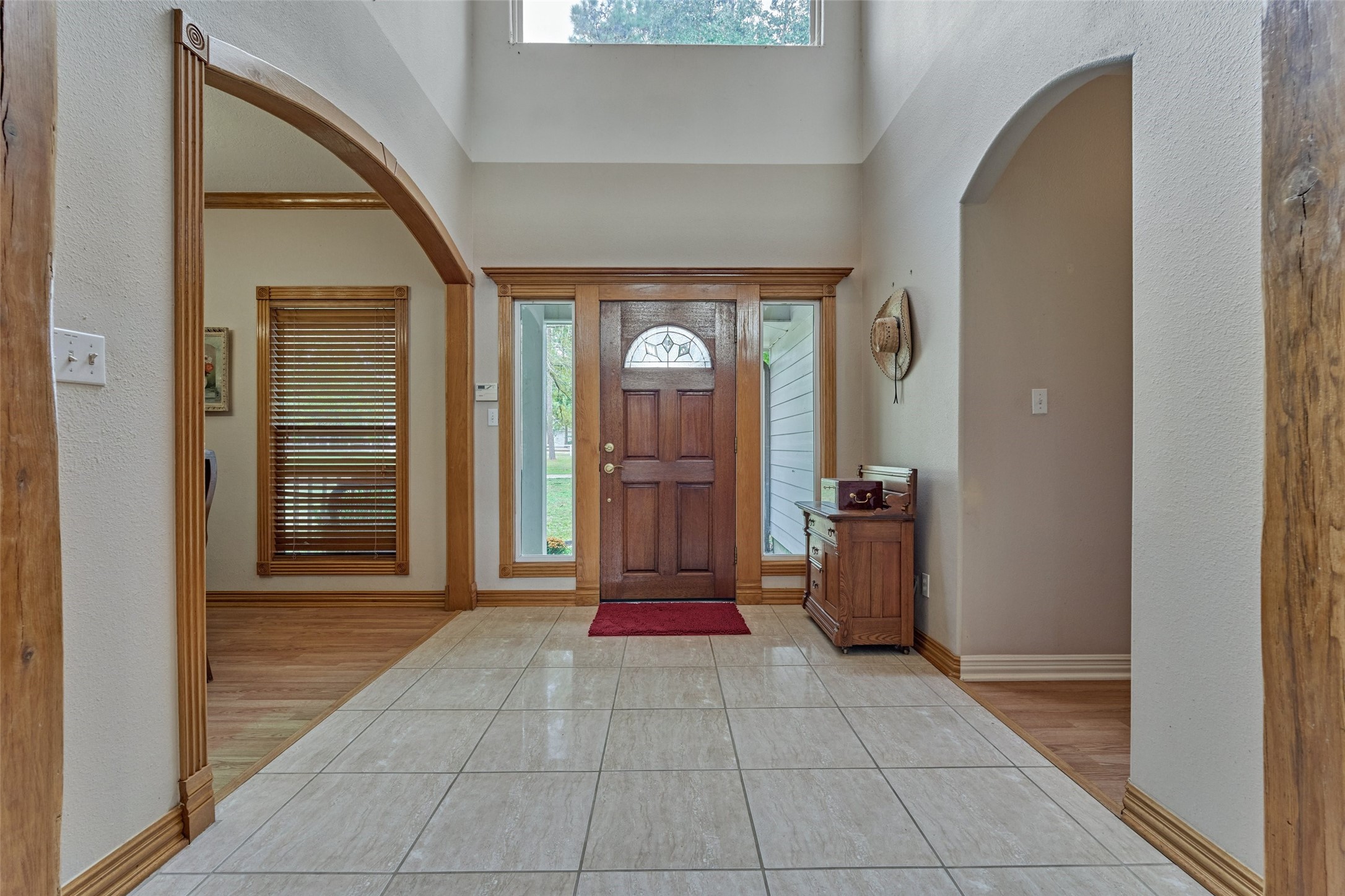 Inviting foyer with high ceilings, a rich wooden front door, and ample natural light, complemented by tile flooring and arched entryways.