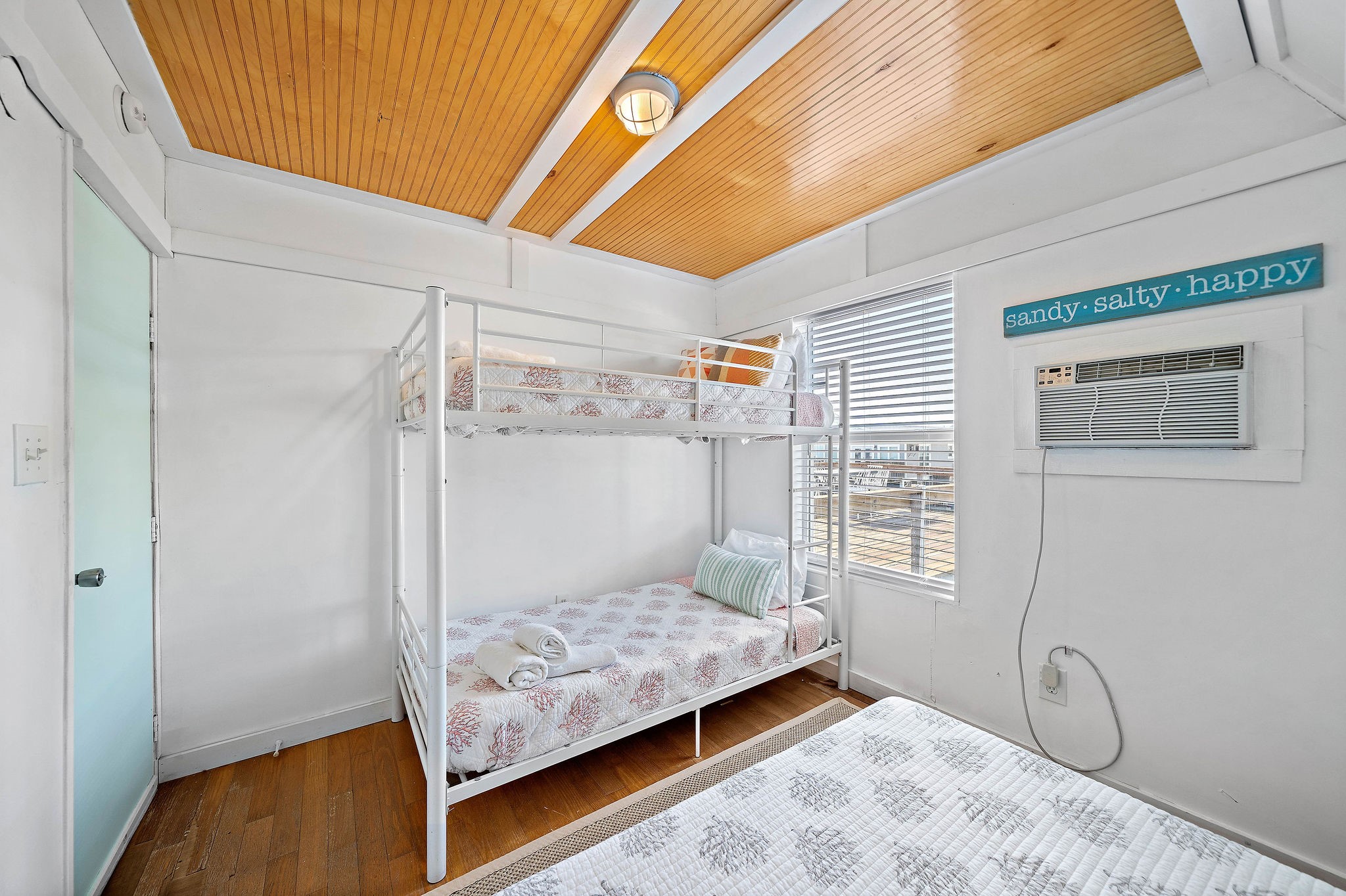 Bright and inviting bedroom featuring a stylish bunk bed, wood-paneled ceiling, and a large window for natural light. A wall-mounted air conditioner ensures comfort, while a cheerful decor sign adds a playful touch.