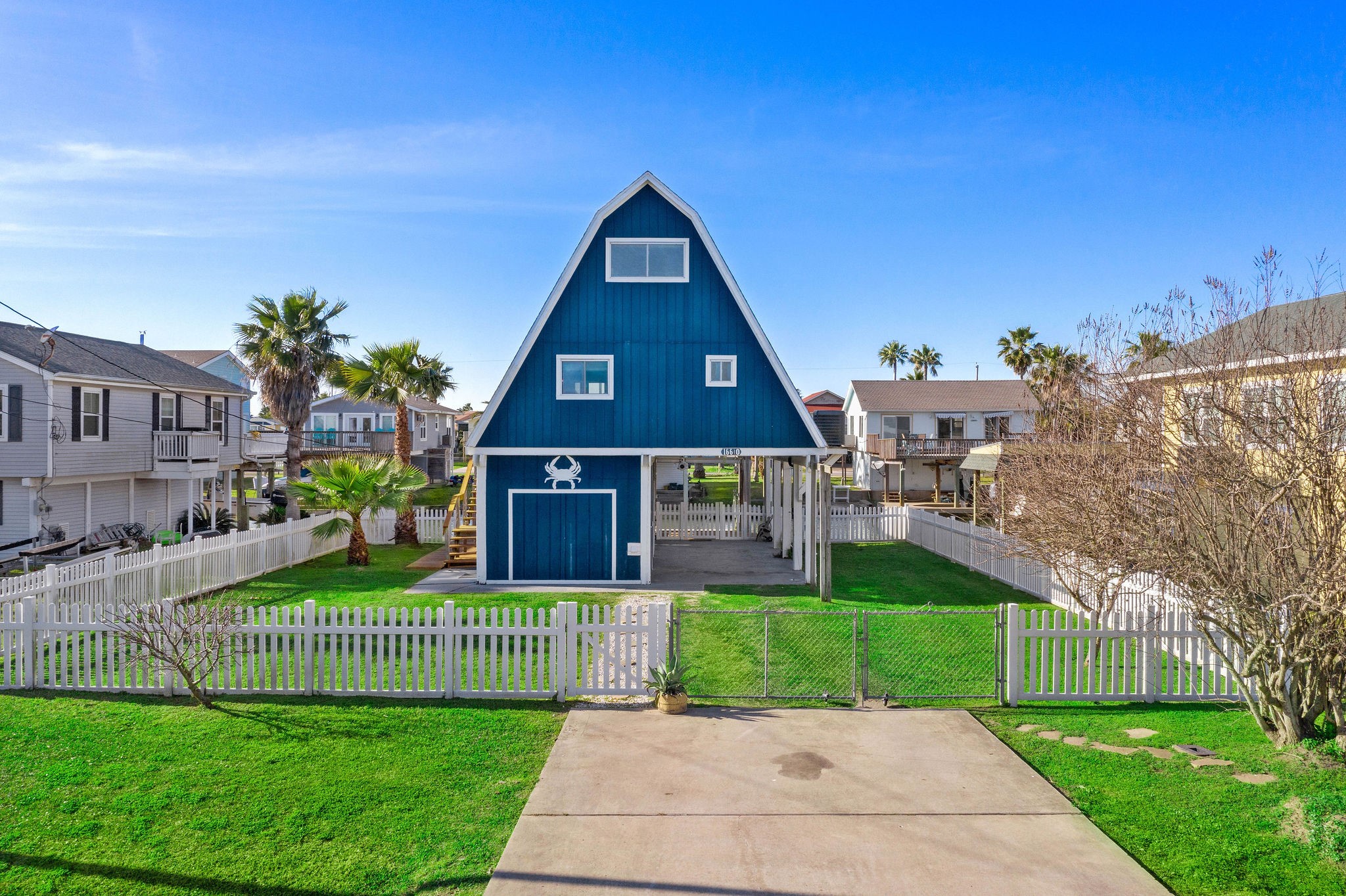Charming A-frame blue house with a white picket fence, spacious driveway, and lush green lawn, offering a coastal feel with palm trees and a welcoming front porch.