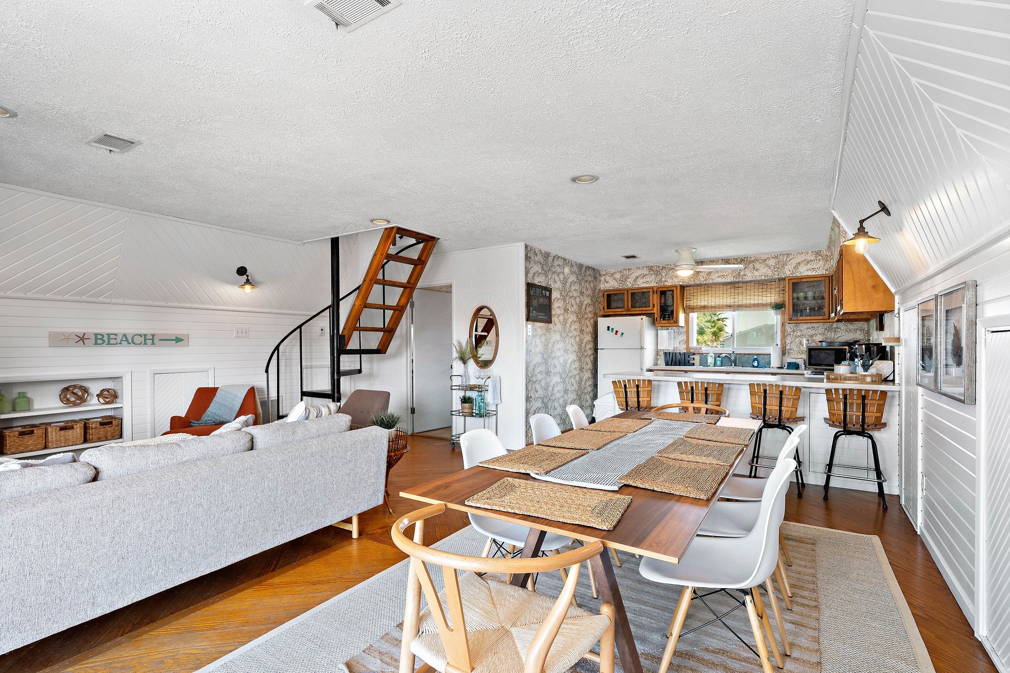 Charming open-concept living space with a cozy seating area, stylish dining table, and a kitchen featuring rustic wood accents. The spiral staircase adds character, and the beach-themed dcor creates a relaxed, inviting atmosphere. Charming open-concept living space with a cozy seating area, stylish dining table, and a kitchen featuring rustic wood accents. The spiral staircase adds character, and the beach-themed dcor creates a relaxed, inviting atmosphere.
