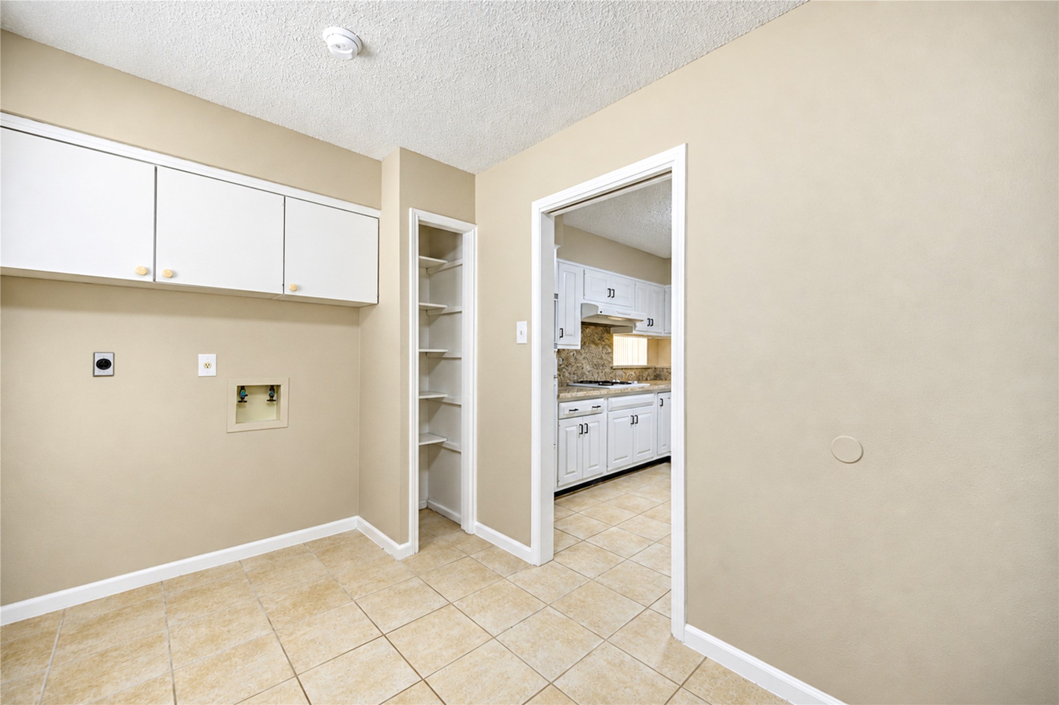 Spacious utility room with tile flooring, built-in cabinets, and a convenient open doorway leading to a modern kitchen with white cabinetry and a granite backsplash.