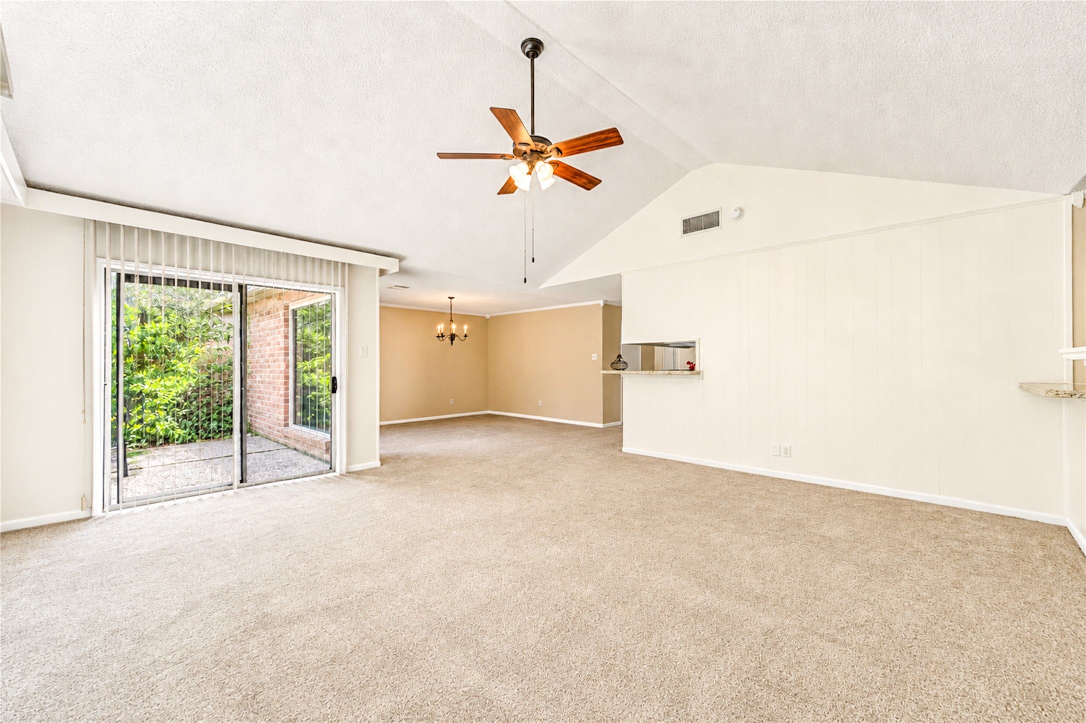 Spacious living area with vaulted ceilings, featuring a ceiling fan and abundant natural light from sliding glass doors leading to a patio.