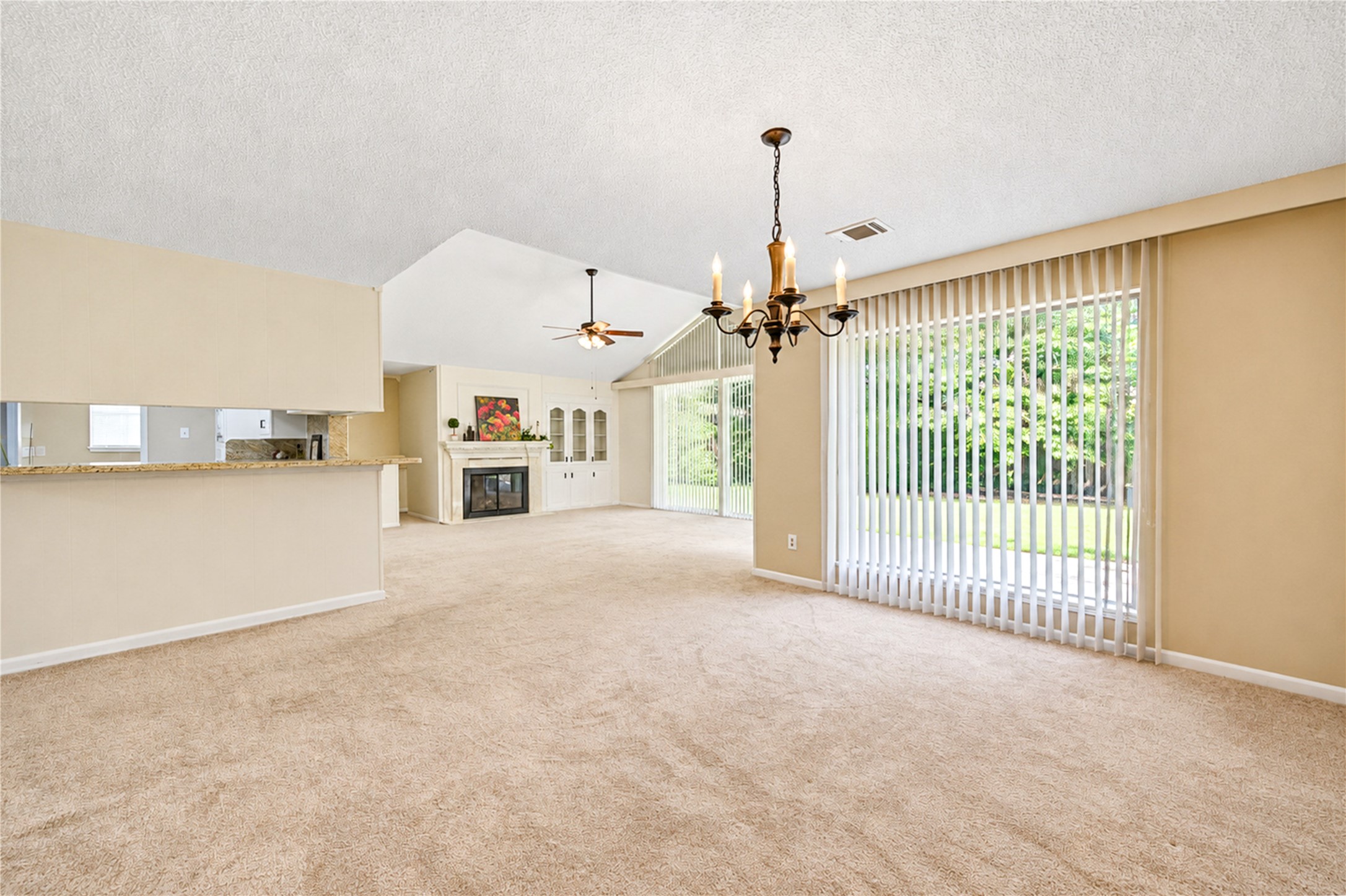 Spacious living area with an open layout, featuring a cozy fireplace, large windows with vertical blinds offering abundant natural light, and a charming chandelier over the dining space.