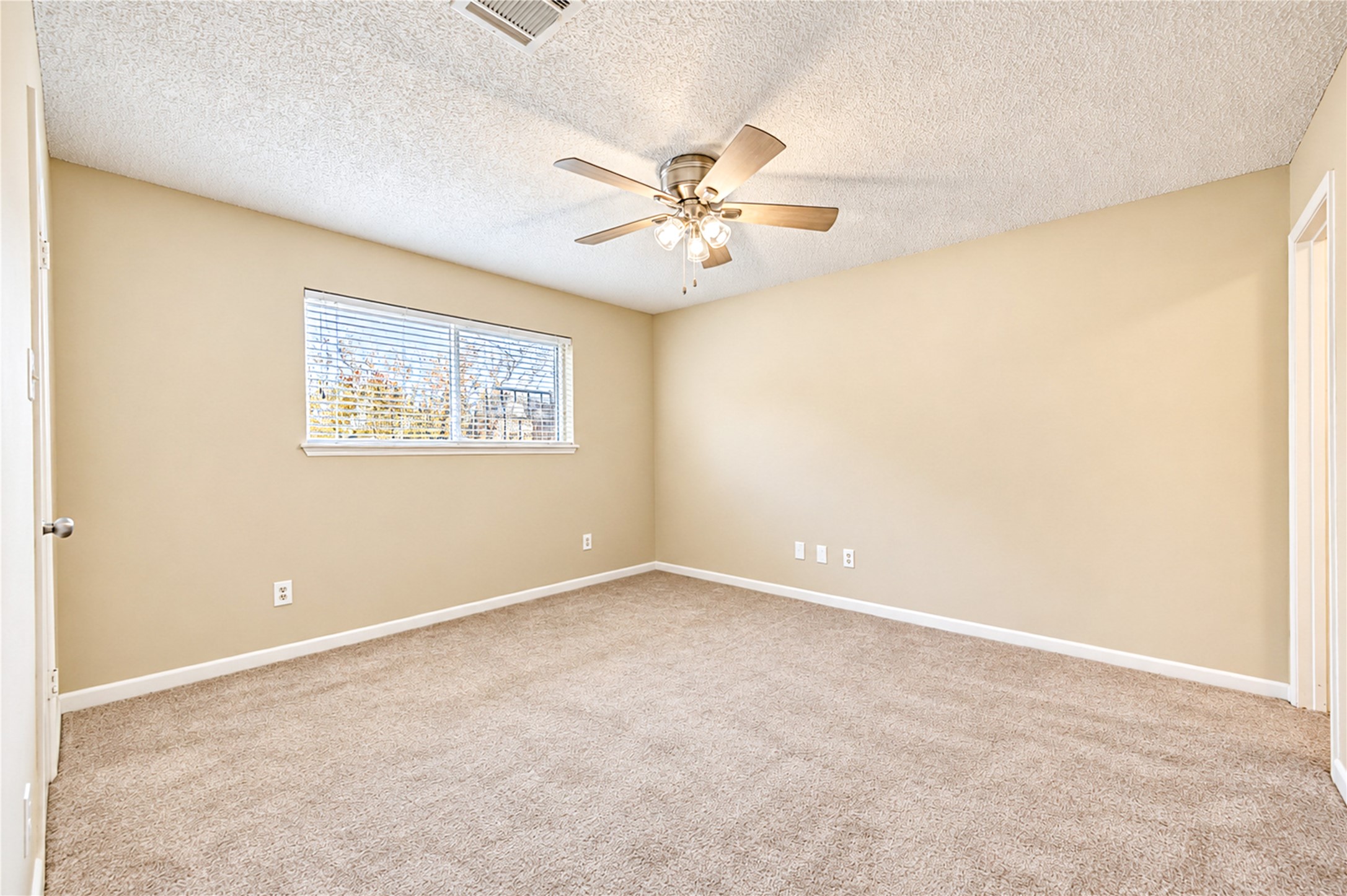 Spacious bedroom with plush carpet, neutral walls, and a large window for natural light, complemented by a stylish ceiling fan.