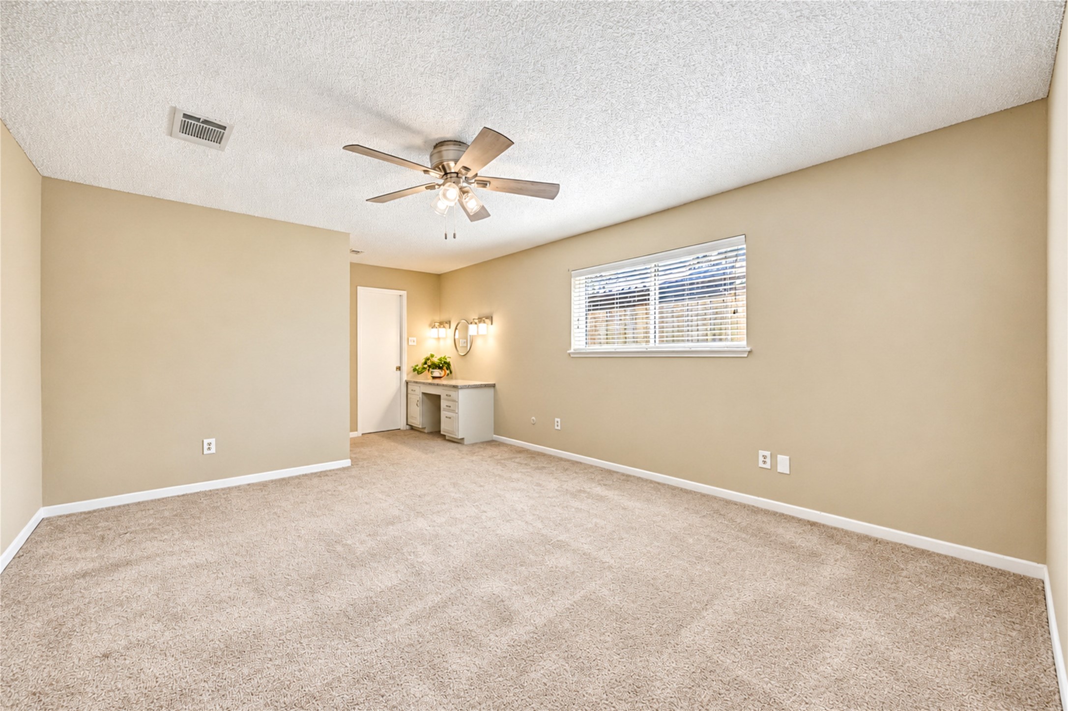 Spacious bedroom with plush carpeting and warm neutral walls, featuring a ceiling fan and a cozy vanity area with natural light from a large window.