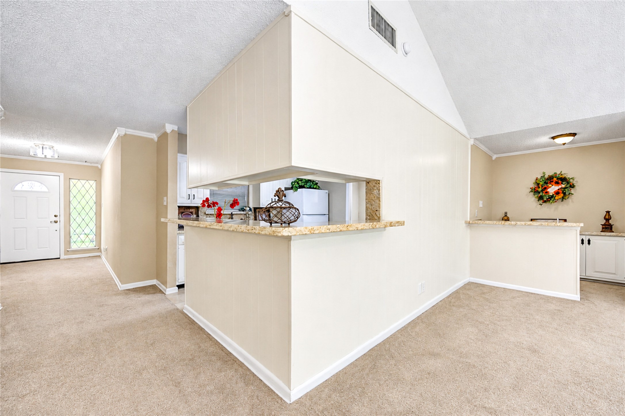 Spacious living area with neutral tones, featuring a cozy carpet, vaulted ceiling, and an open pass-through to the kitchen. The entryway is inviting with a decorative glass-panel door and ample natural light.