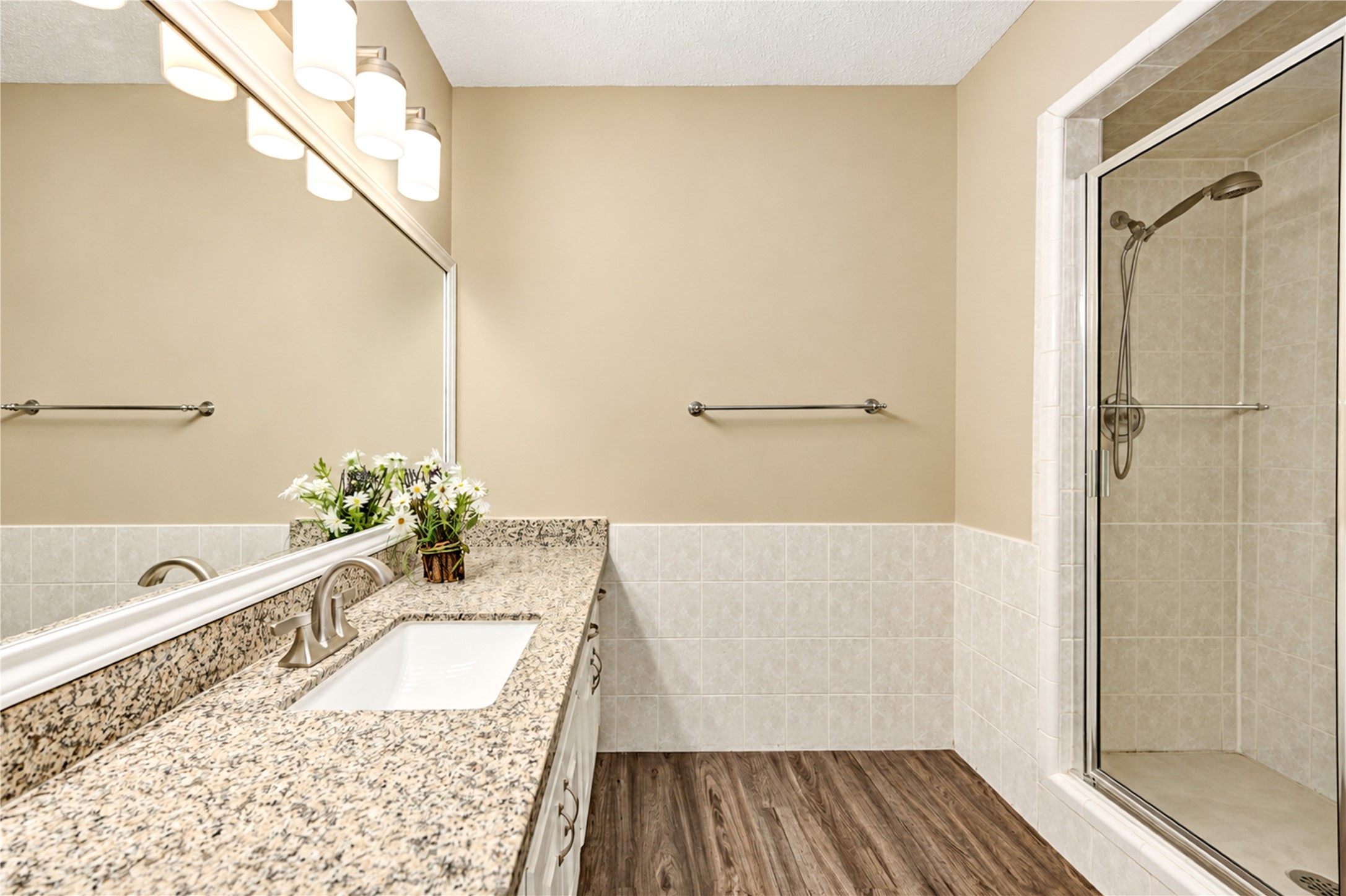 Elegant primary bedroom with ensuite and  a spacious granite countertop with under mount sink, modern fixtures, and a glass-enclosed shower, complemented by warm lighting and wood-style flooring.