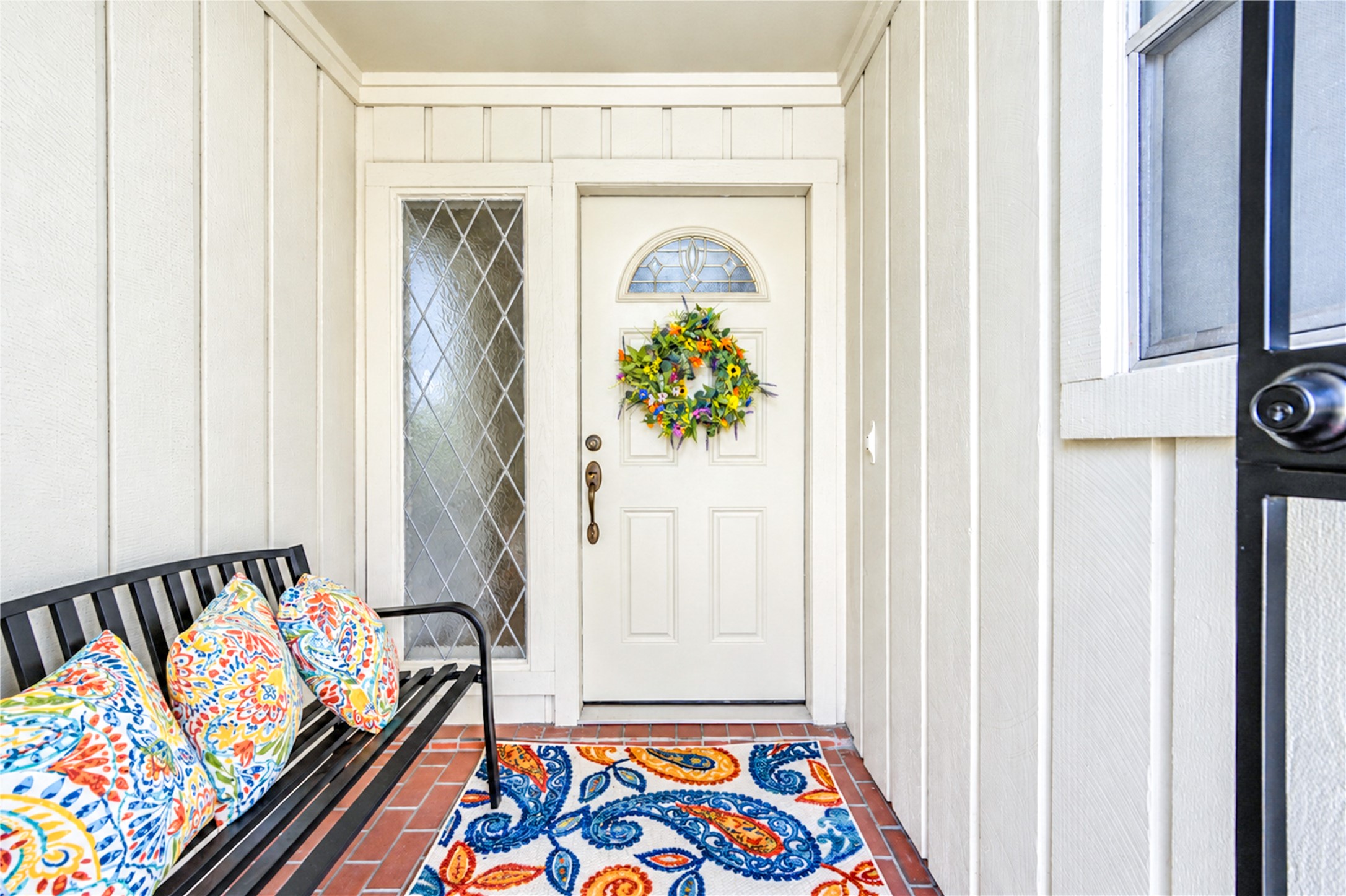 Charming entryway featuring a white door with an elegant decorative glass panel, complemented by a vibrant wreath and colorful cushions on a sleek bench.