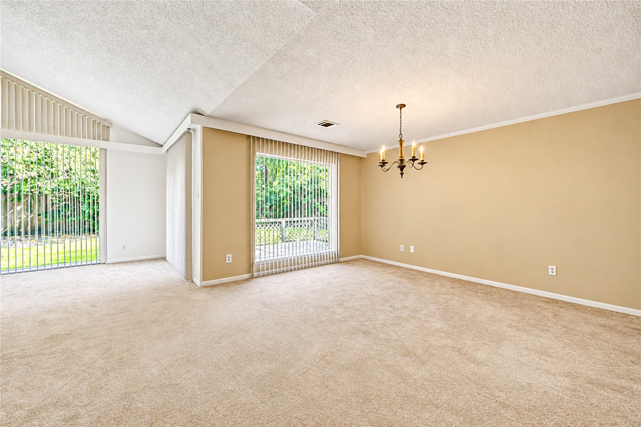 Spacious living area with plush carpeting, a stylish chandelier, and large windows offering abundant natural light and garden views.