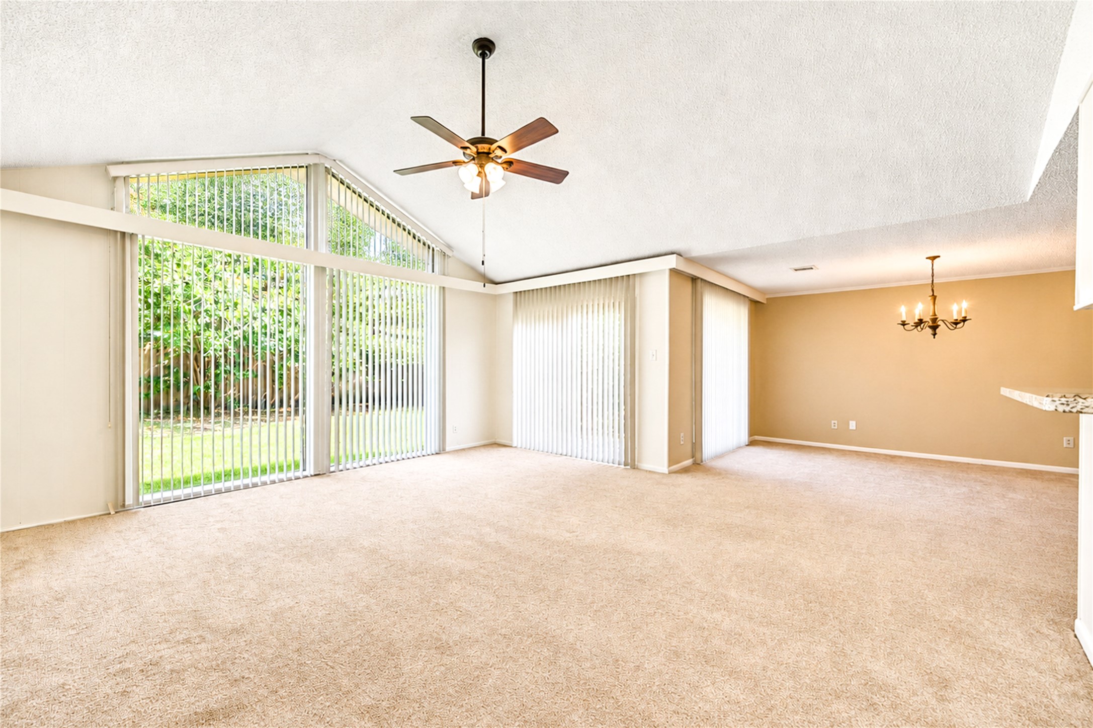 Spacious living area with vaulted ceilings and abundant natural light from large windows, complemented by a cozy carpeted floor and elegant chandelier.