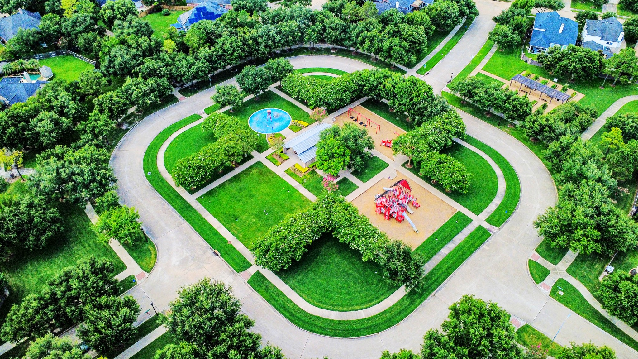 This aerial photo showcases a vibrant neighborhood park surrounded by a circular street. The park features a playground, open green spaces, and a splash pad, providing a family-friendly environment. Lush trees line the streets, and well-maintained homes are visible around the perimeter, indicating a tranquil and inviting residential area.