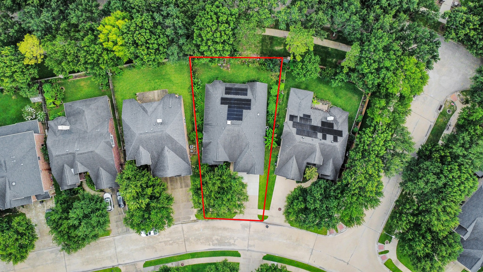 This aerial view shows a house with a spacious backyard outlined in red, nestled in a leafy neighborhood. The roof has solar panels, and the property is surrounded by mature trees, offering privacy and greenery.