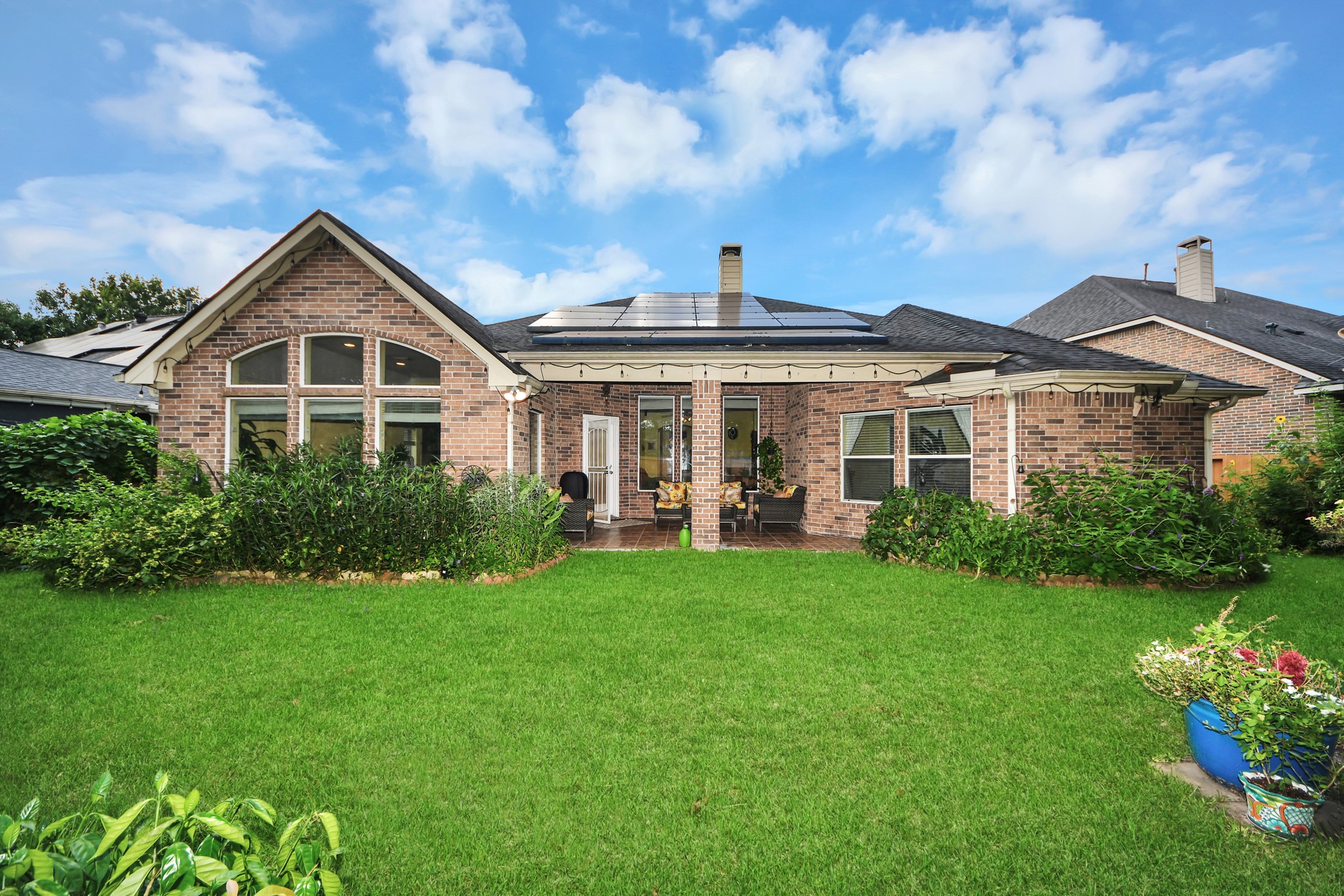 This photo showcases a charming brick home with solar panels on the roof. It features a well-maintained backyard with lush greenery and a cozy patio area, perfect for outdoor relaxation.