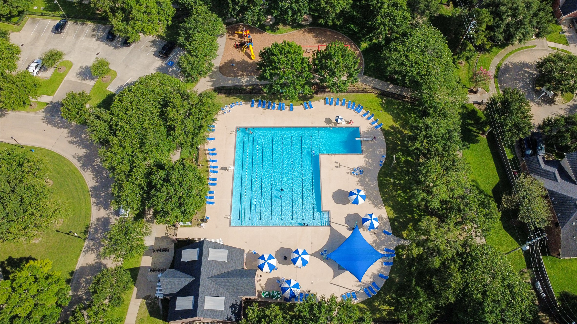 This aerial view showcases a well-maintained community pool area surrounded by lush greenery. It features a large swimming pool, ample lounge chairs, umbrellas, a shaded pavilion, and a nearby playground, offering a perfect recreational spot for residents.