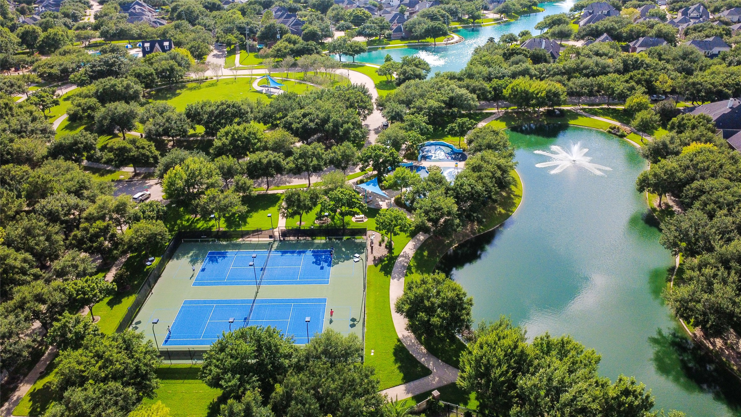 This aerial photo showcases a picturesque residential community featuring lush greenery, two tennis courts, winding walking paths, a serene pond with a fountain, and a swimming pool. Ideal for active lifestyles and relaxation.