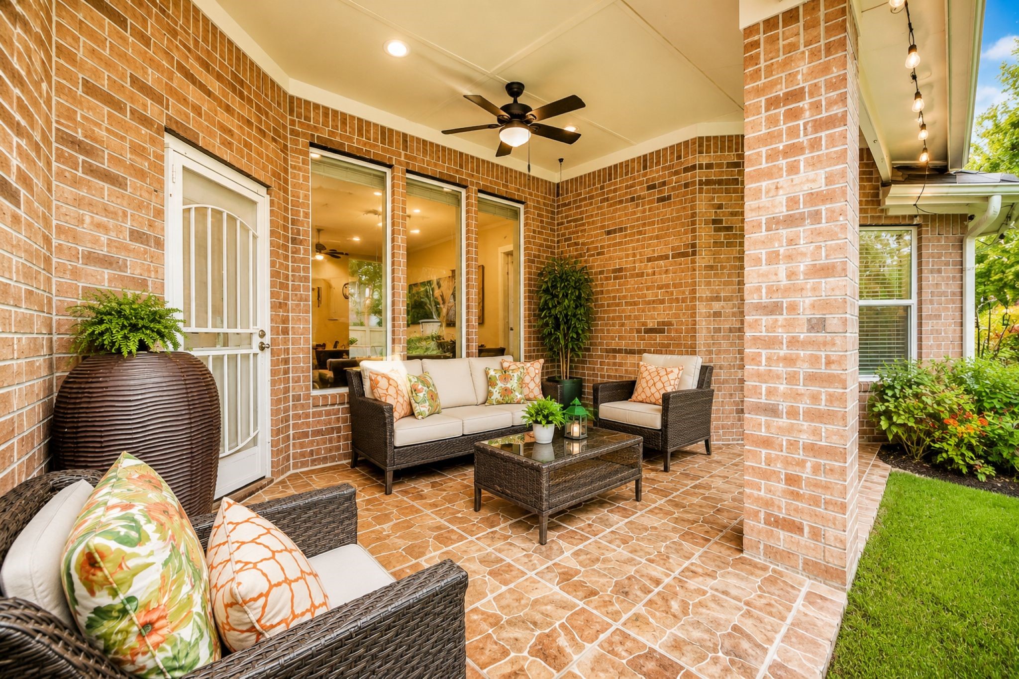 VIRTUALLY STAGED!
This inviting patio features a cozy seating area with wicker furniture and colorful cushions, set against a backdrop of red brick walls. It has elegant tile flooring, a ceiling fan for comfort, and lush greenery, creating a perfect outdoor relaxation space.