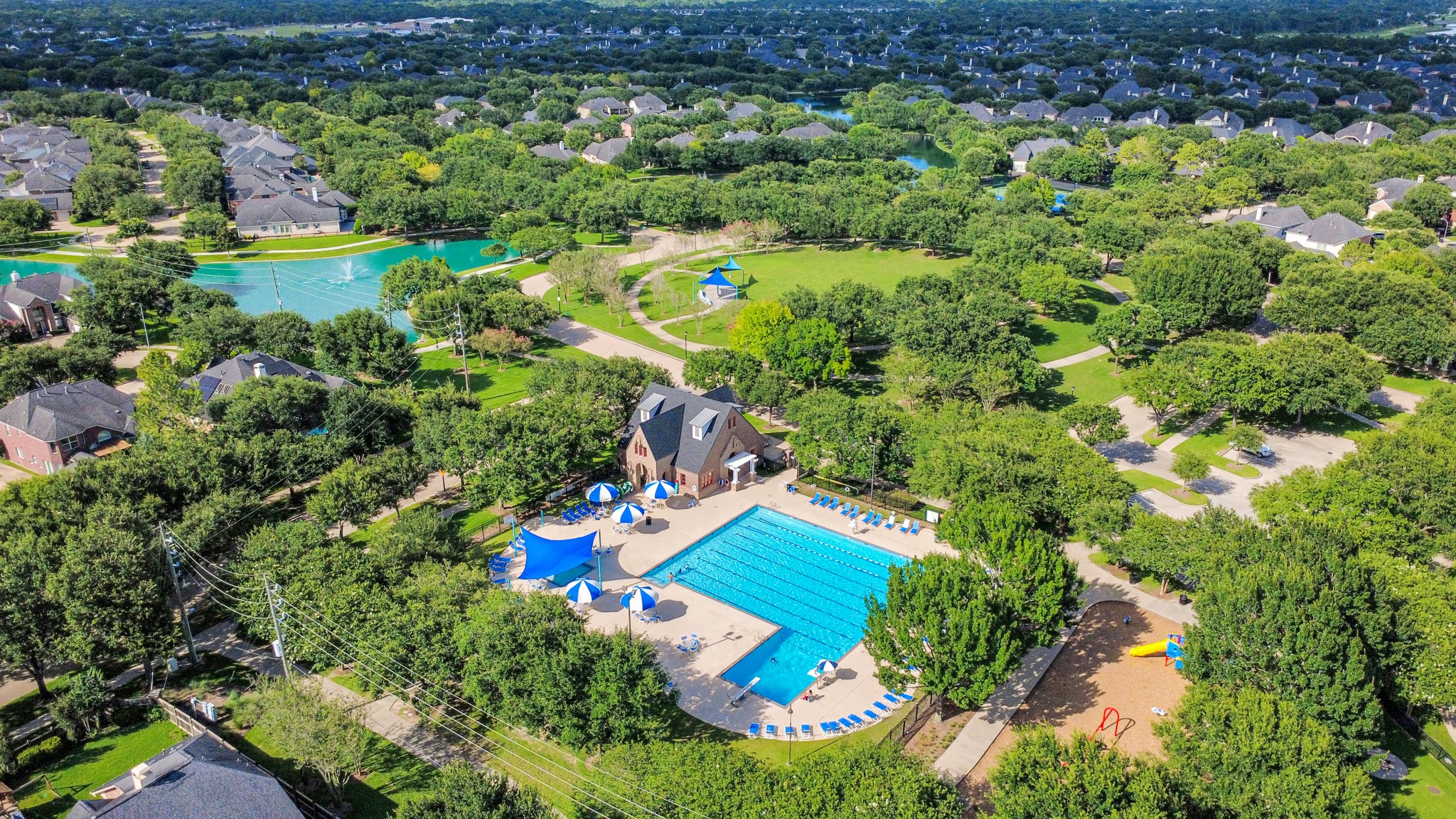 This aerial photo showcases a vibrant neighborhood with lush greenery, featuring a large community pool, playground, and walking paths. Residences are nestled among trees, offering a serene suburban setting.