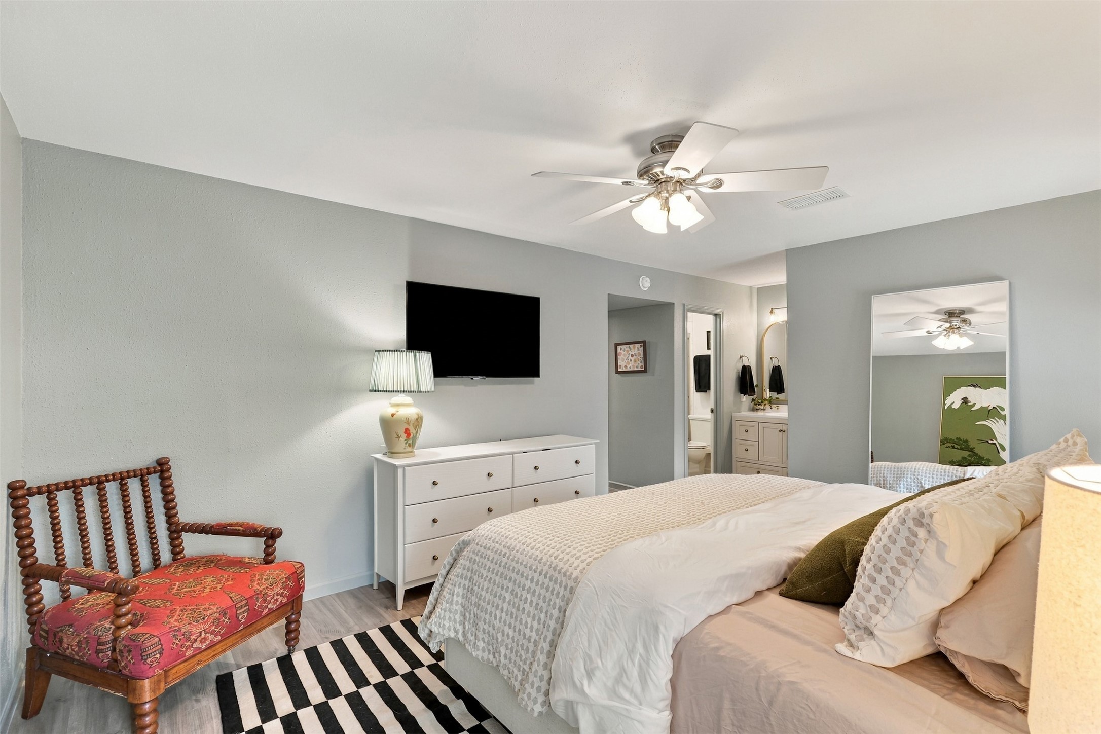 Spacious bedroom with soft gray walls, featuring a cozy bed, a stylish chair, and a sleek dresser topped with a TV. A ceiling fan and ample lighting create a bright, welcoming atmosphere.