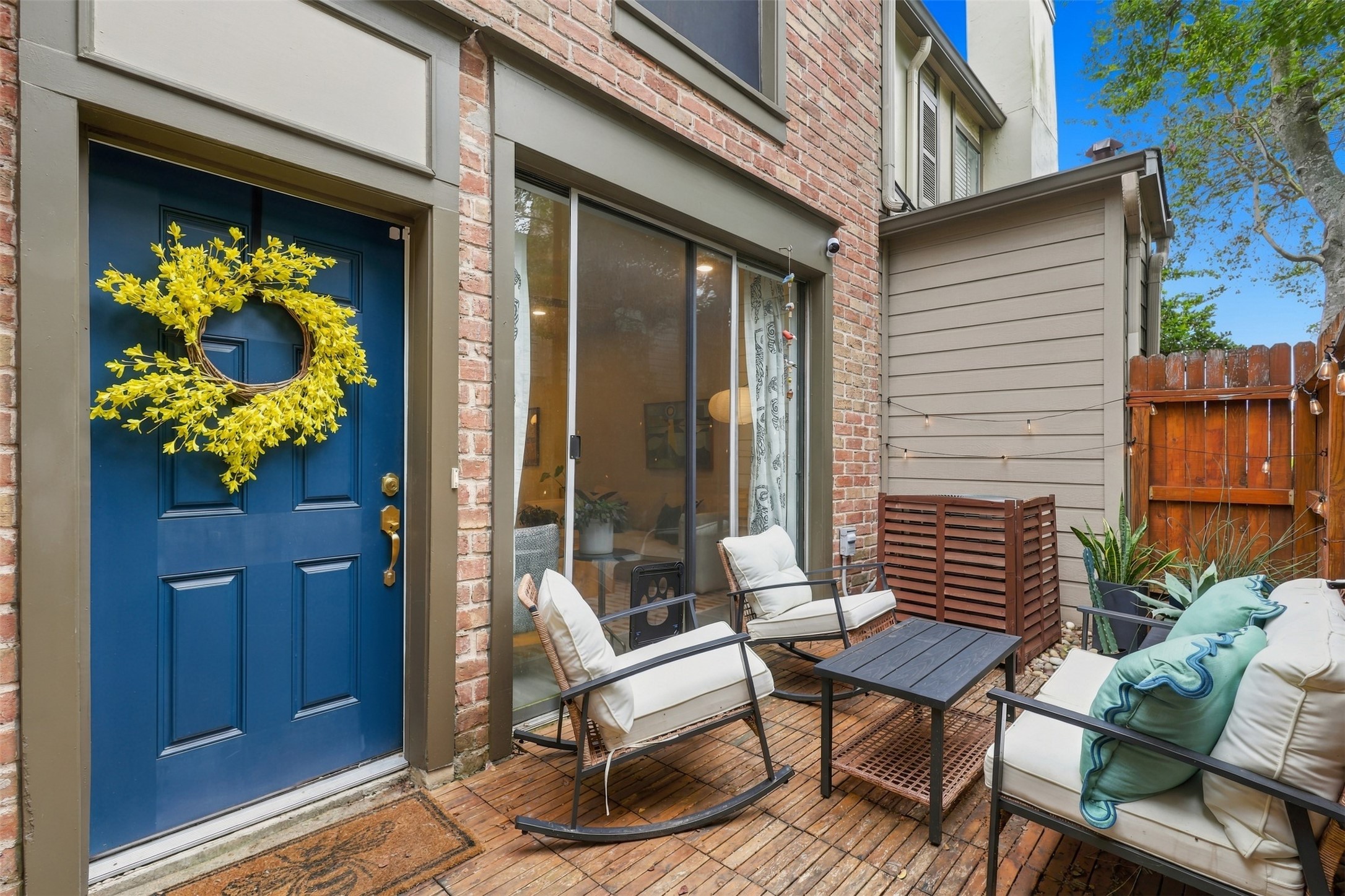 Charming patio features cozy seating, a vibrant blue door with a cheerful wreath, and a warm brick exterior, perfect for relaxing or entertaining.