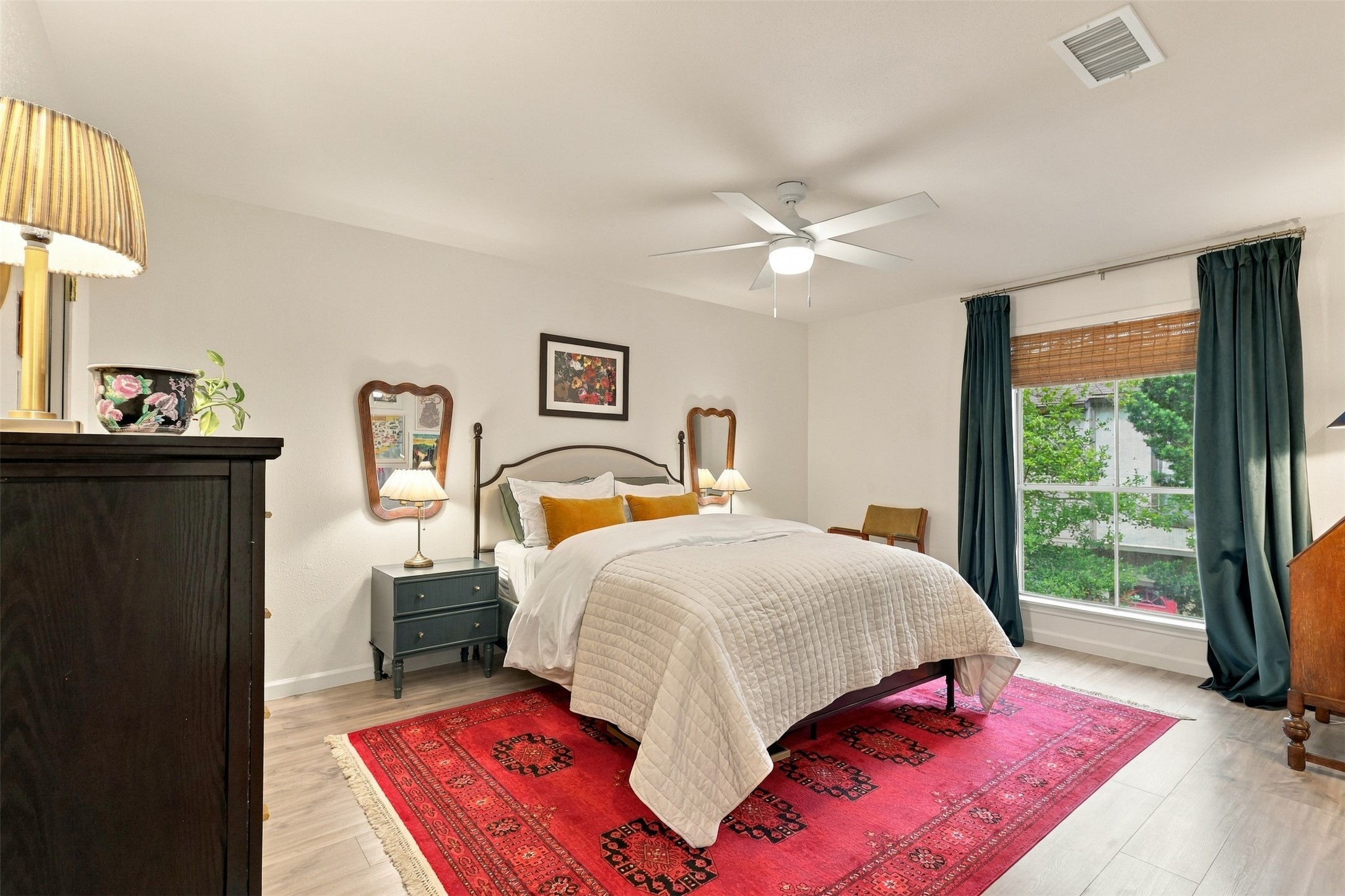 Spacious and bright bedroom featuring a cozy bed with elegant bedding, large window with rich drapes, and a vibrant red area rug adding warmth and style.
