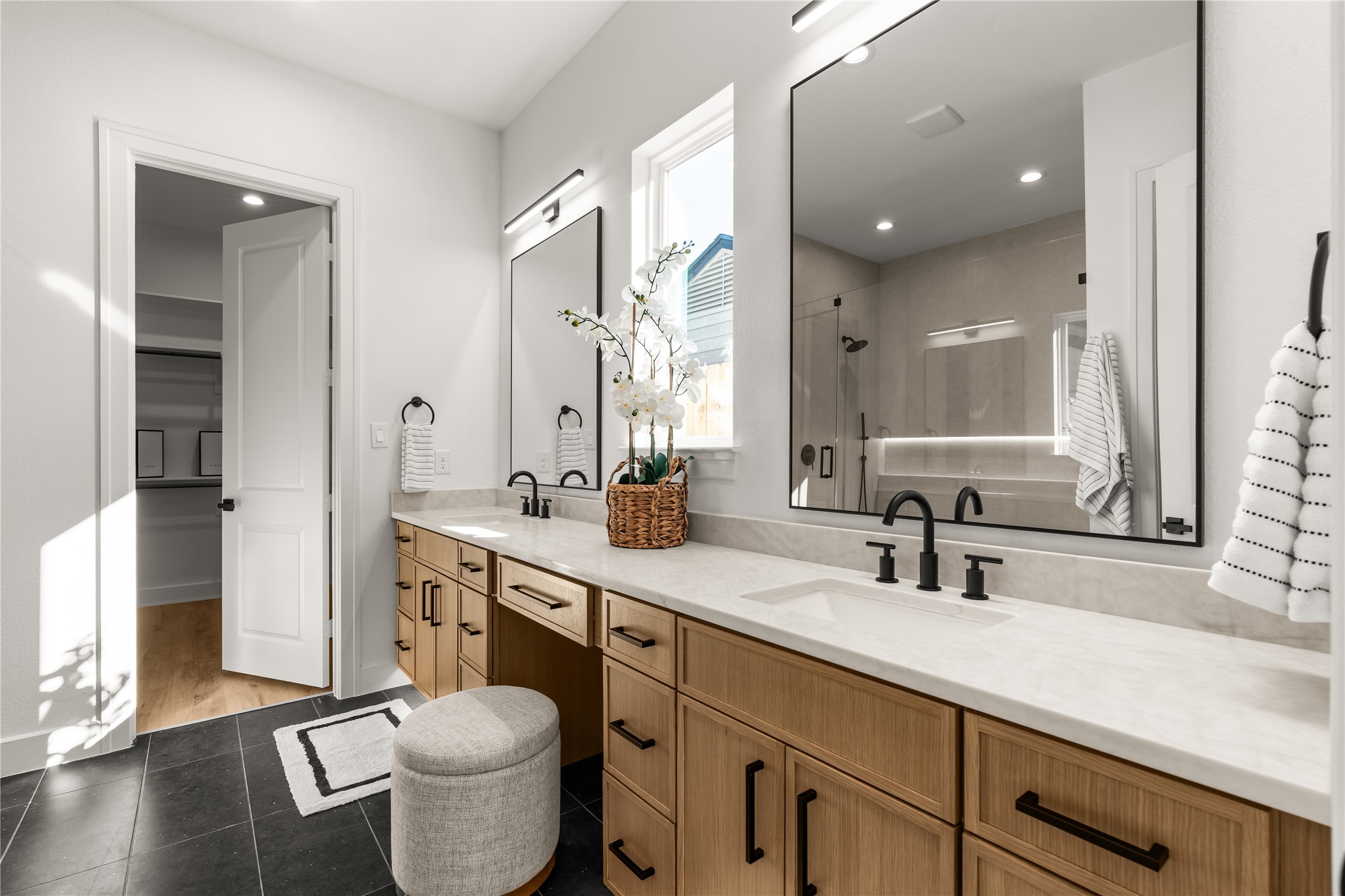 The primary ensuite showcases an expansive dual-sink vanity with warm wood-toned cabinetry, light stone-look countertops, and matte black fixtures.