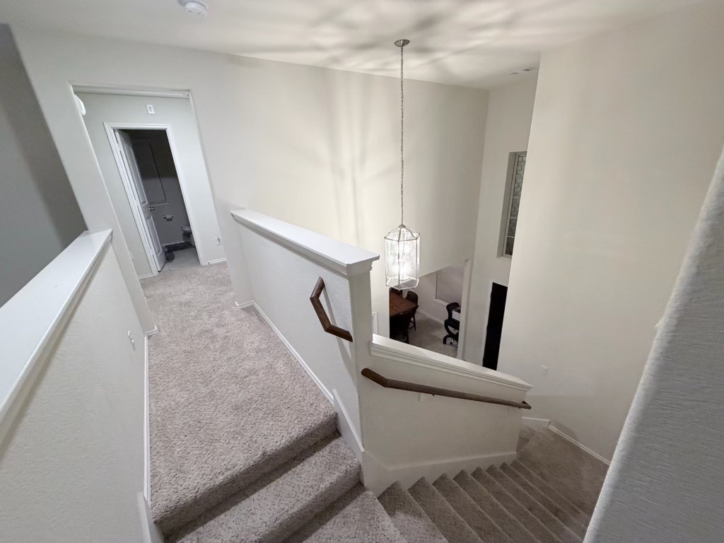 This photo shows a carpeted staircase with a wooden handrail leading down to a dining area. The space is well-lit by a modern hanging light fixture, and there's a hallway with a visible doorway on the upper level. The walls are painted a neutral color, enhancing the bright and open feel.