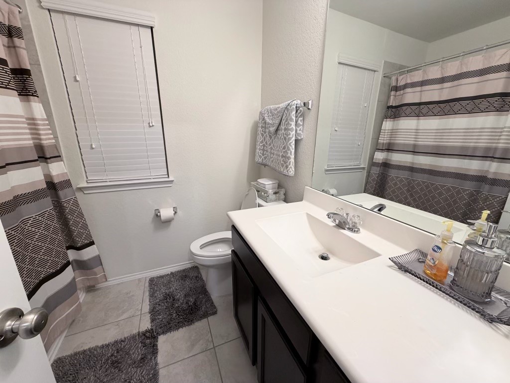 This bathroom features a clean and modern design with neutral tones. It includes a shower-tub combo with a striped curtain, a spacious vanity with a large mirror, and tiled flooring. The window provides natural light, and there are tasteful accessories for a cozy feel.