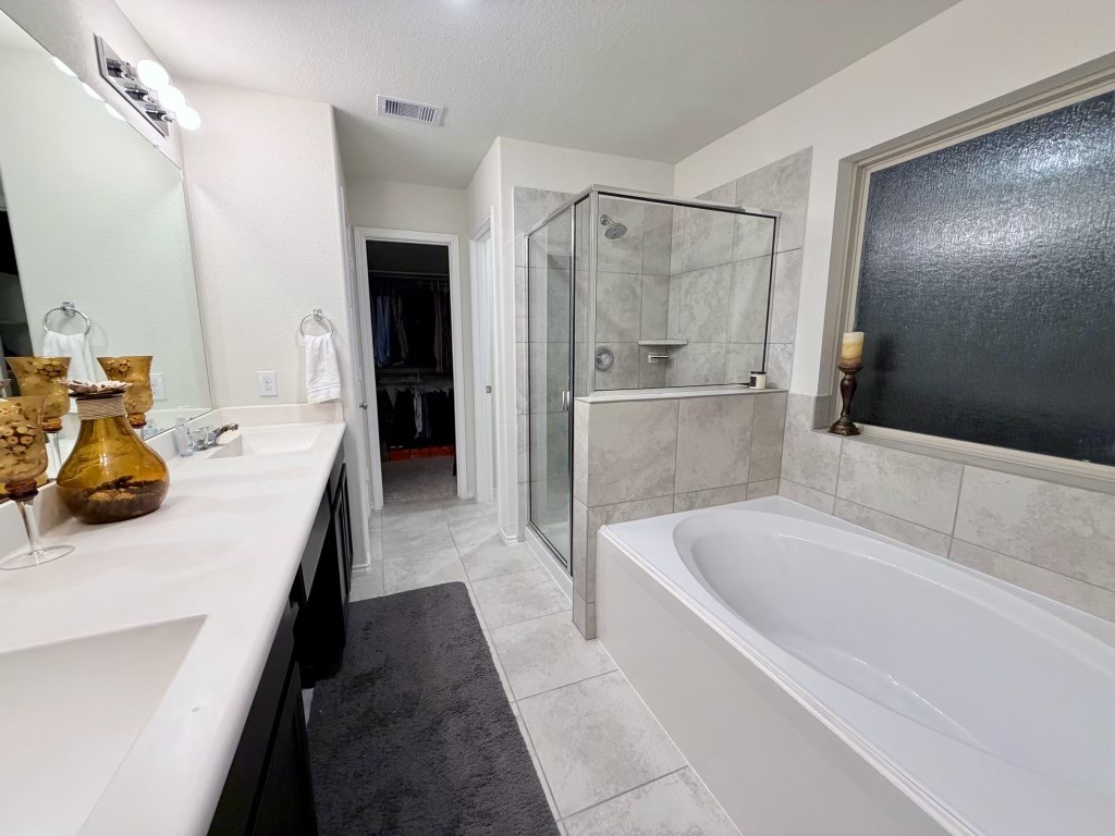 This bathroom features a sleek design with a large vanity, double sink, and modern fixtures. It includes a spacious glass-enclosed shower and a separate soaking tub, complemented by neutral tile and a frosted window for privacy.