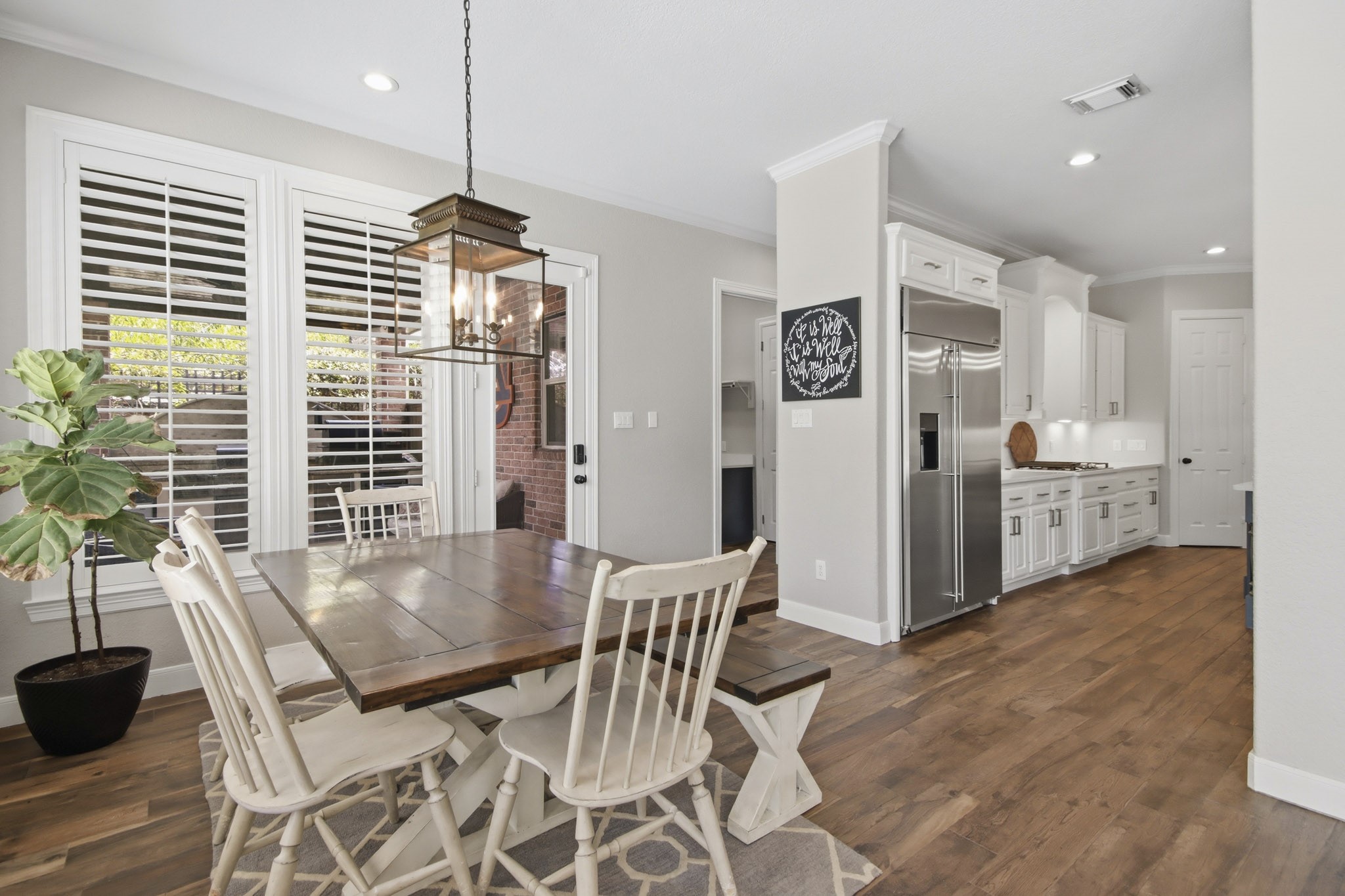 Bright and inviting breakfast area with elegant light fixture, and large windows with shutters, seamlessly connected to kitchen and living room.