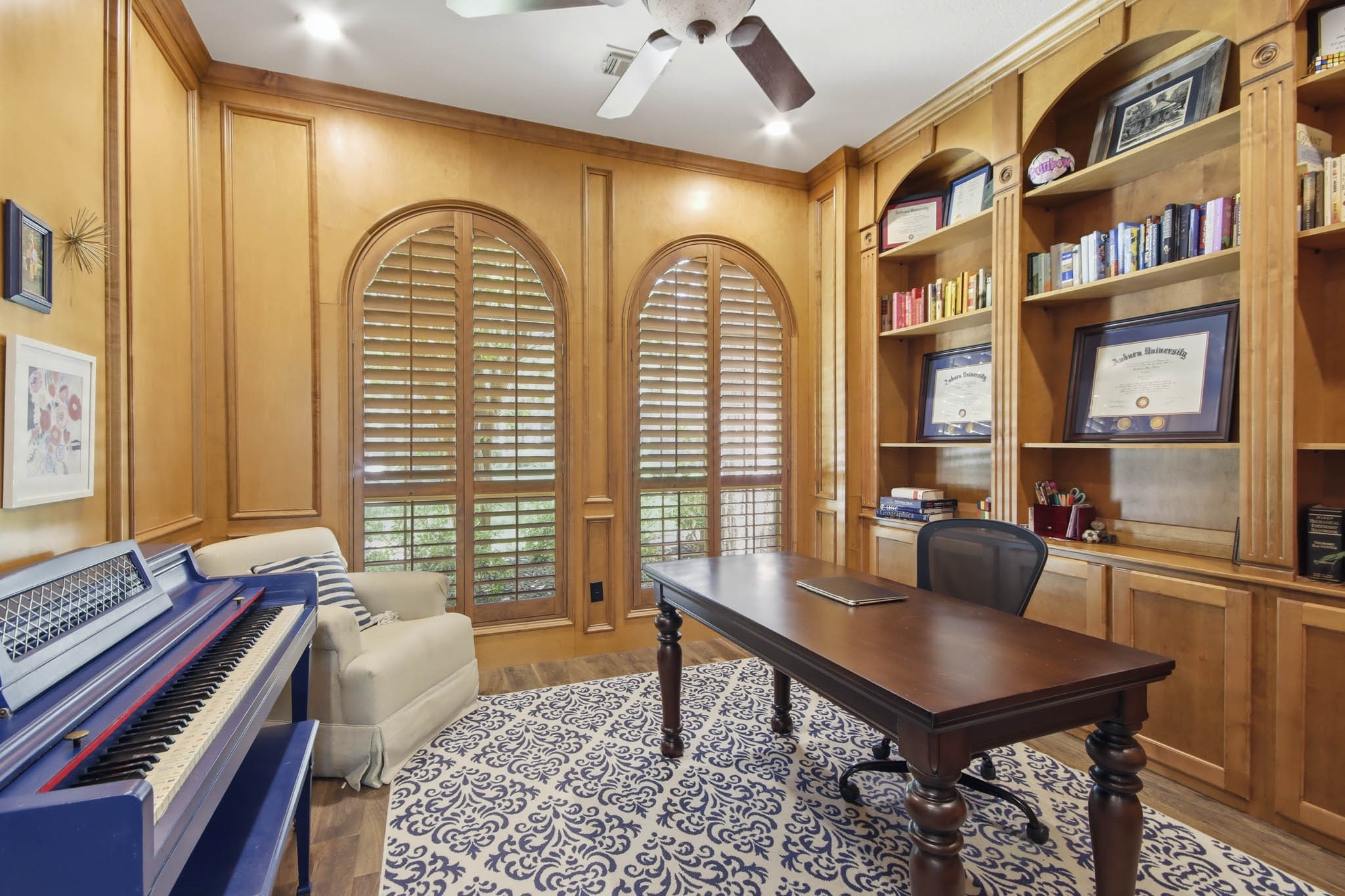 Home office featuring rich wood paneling, custom built-in bookshelves, and arched windows with shutters creating a perfect space for productivity and inspiration.