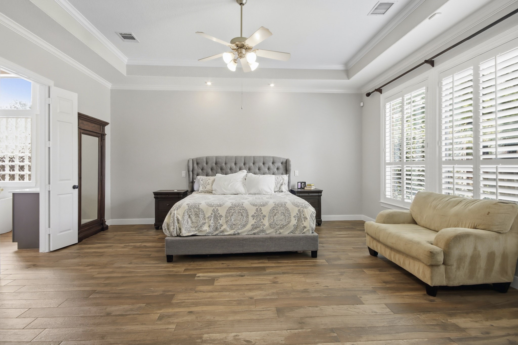 Spacious bedroom with wood-like tile floors, ample natural light streaming through large shuttered windows, complemented by enough space for a cozy seating area.