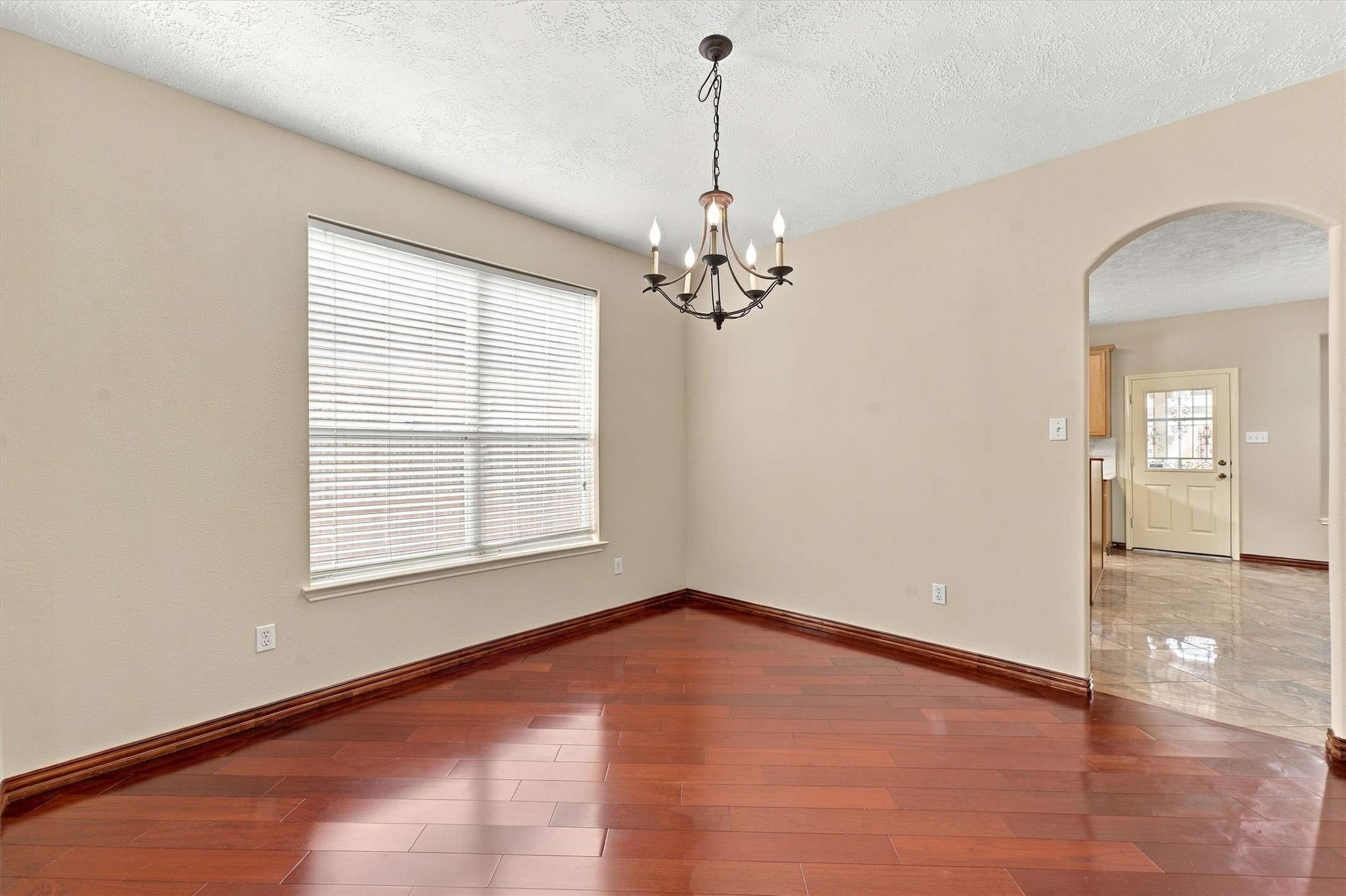 Elegant dining room with rich hardwood floors, a classic chandelier, and ample natural light, leading to a tiled kitchen area through a graceful archway.