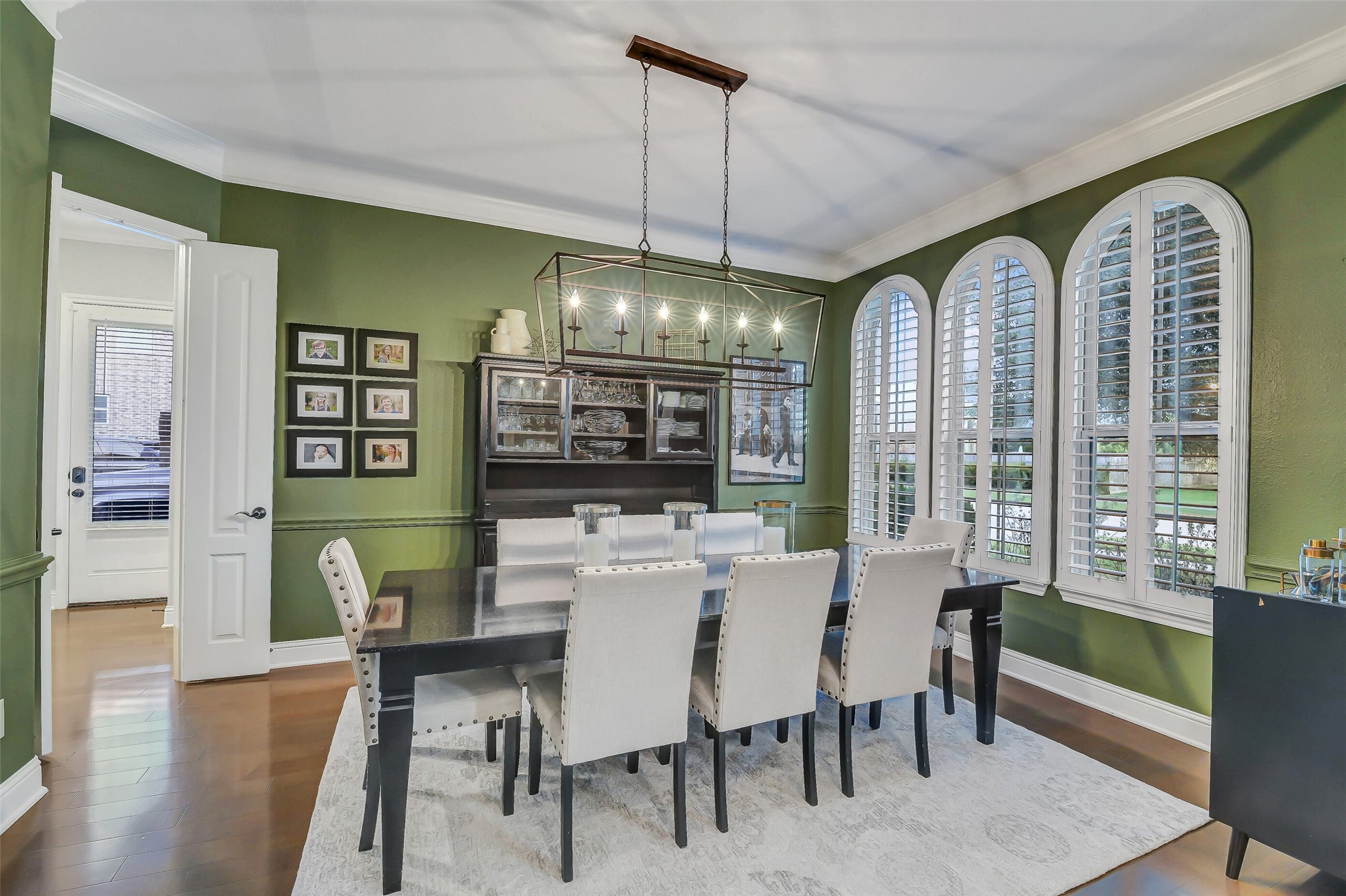 An elegant formal dining room highlighted by a stunning wall of arched windows with plantation shutters, hardwood flooring, crown molding, and a statement chandeliercreating the perfect setting for memorable gatherings and special occasions