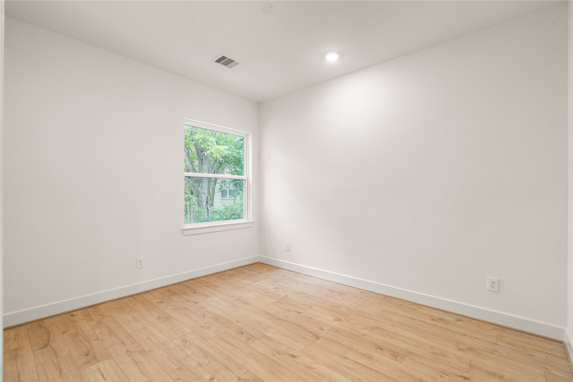 Quiet corner bedroom  warm oak-look flooring, window overlooking mature greenery, and clean neutral paint finishes.