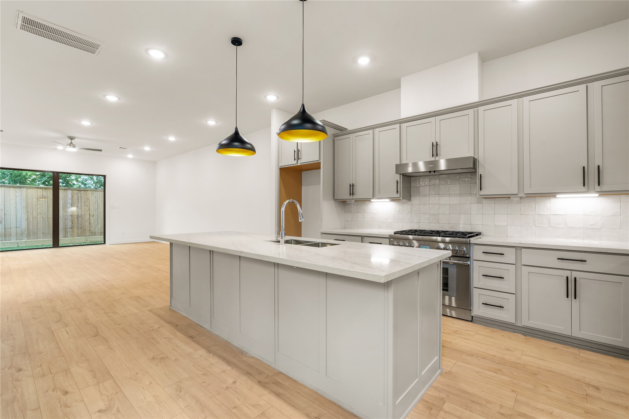 Chef's kitchen perspective  massive quartz island with bar seating potential, statement matte black and gold pendant lighting, gas cooktop setup, and soft grey shaker cabinetry extending to the main floor living space.