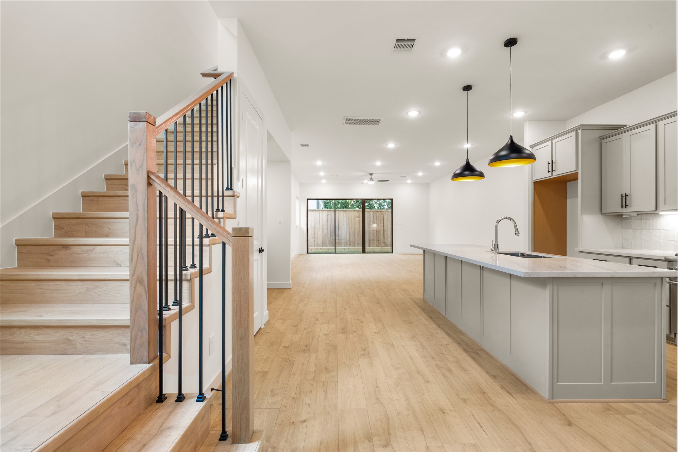 Grand main-level floor plan  expansive quartz kitchen island, matte black dome pendant lights, custom wood-and-iron staircase, and floor-to-ceiling glass patio doors.