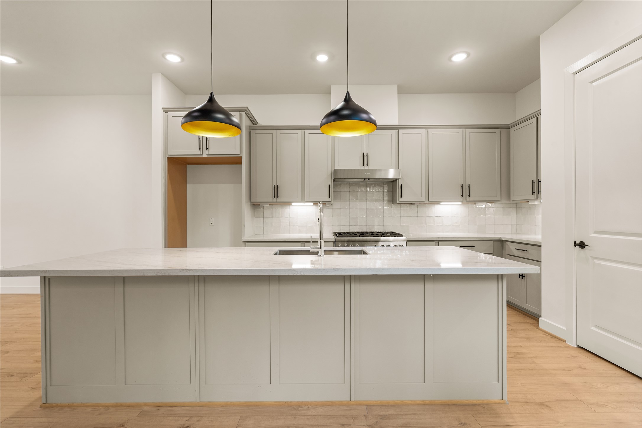 The culinary center  expansive quartz prep island with undermount double sink, stainless steel range hood, under-cabinet lighting, and clean tile backsplash.