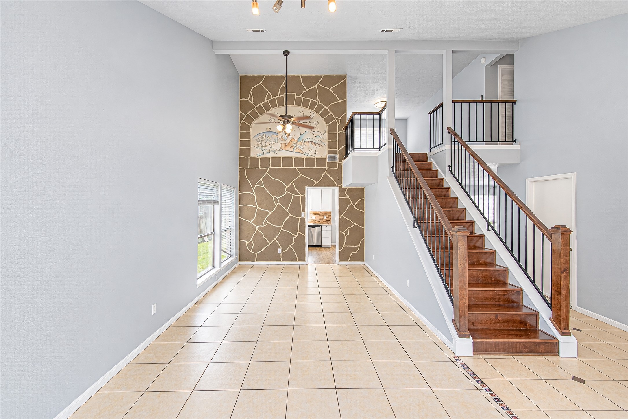 Spacious open living area with elegant tile flooring, a striking stone accent wall, and a grand staircase leading to the upper level, exuding warmth and style. Spacious open living area with elegant tile flooring, a striking stone accent wall, and a grand staircase leading to the upper level, exuding warmth and style.