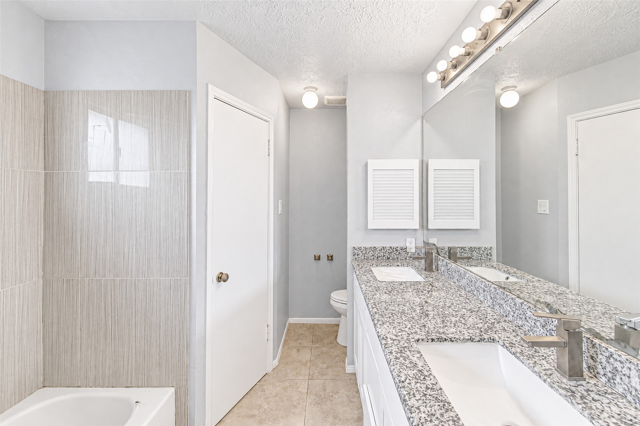 Bright and modern bathroom featuring dual sinks with sleek granite countertops, a large mirror, and stylish light fixtures. Neutral tile surrounds the bathtub, creating a clean and inviting atmosphere.
