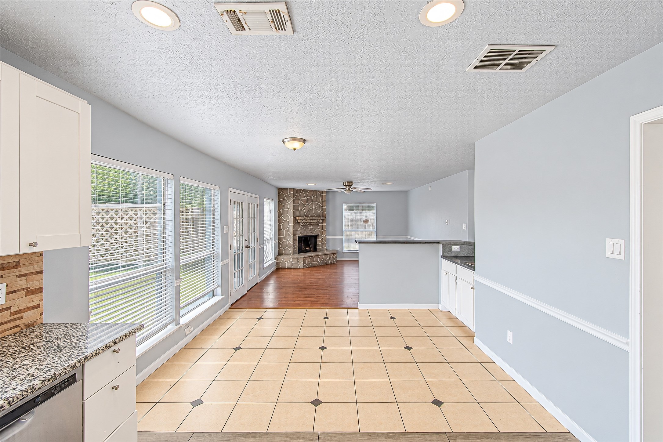 Bright and open living space with large windows, featuring a cozy stone fireplace and a seamless transition from tiled kitchen to hardwood floors.