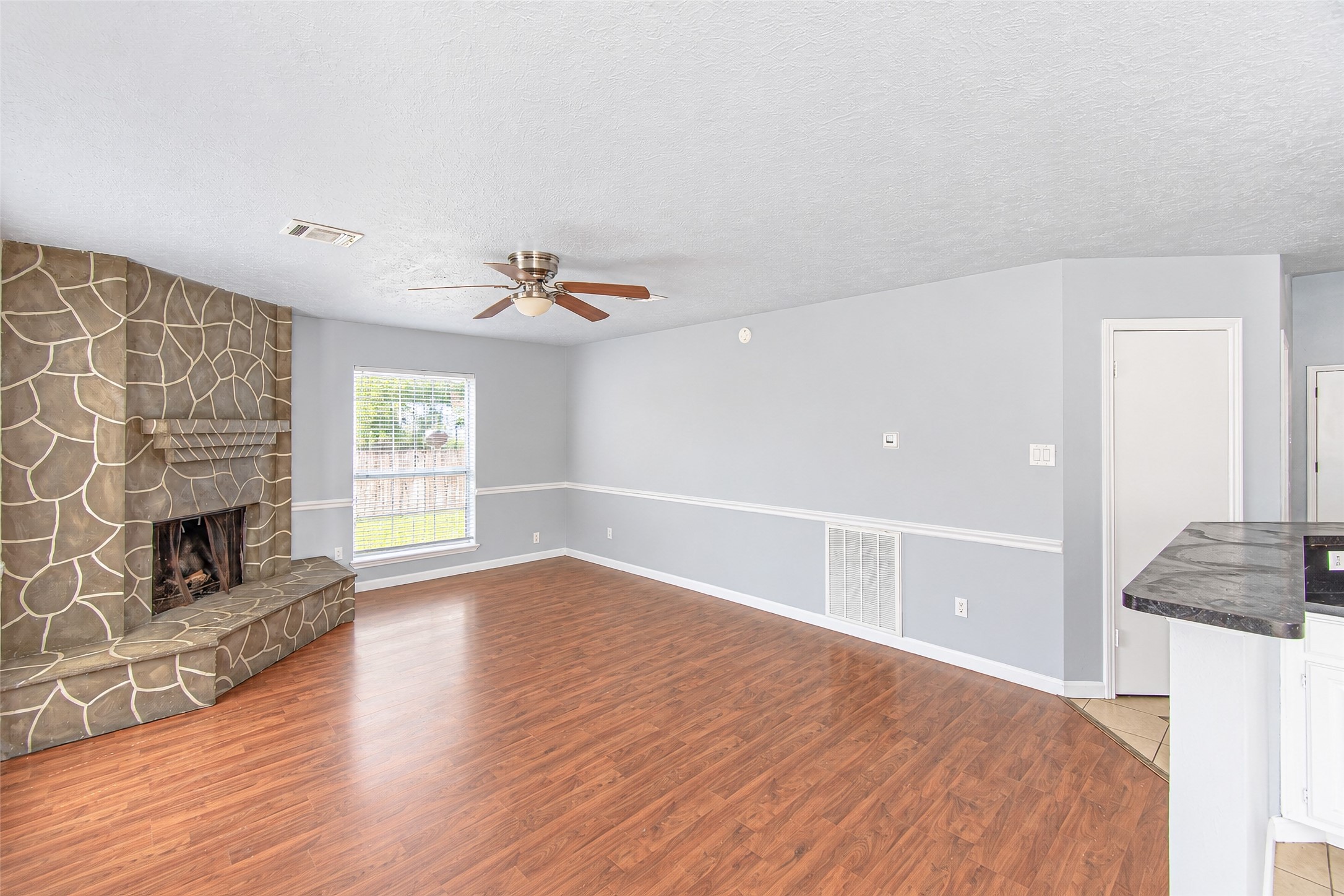 Spacious living area with elegant wood flooring and a unique stone fireplace, complemented by soft gray walls and ample natural light.