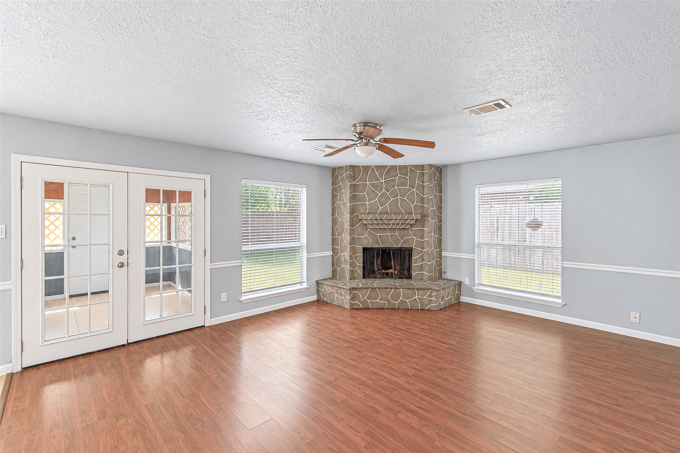 Spacious living room with elegant wood flooring, a striking stone fireplace, and abundant natural light from large windows and French doors.