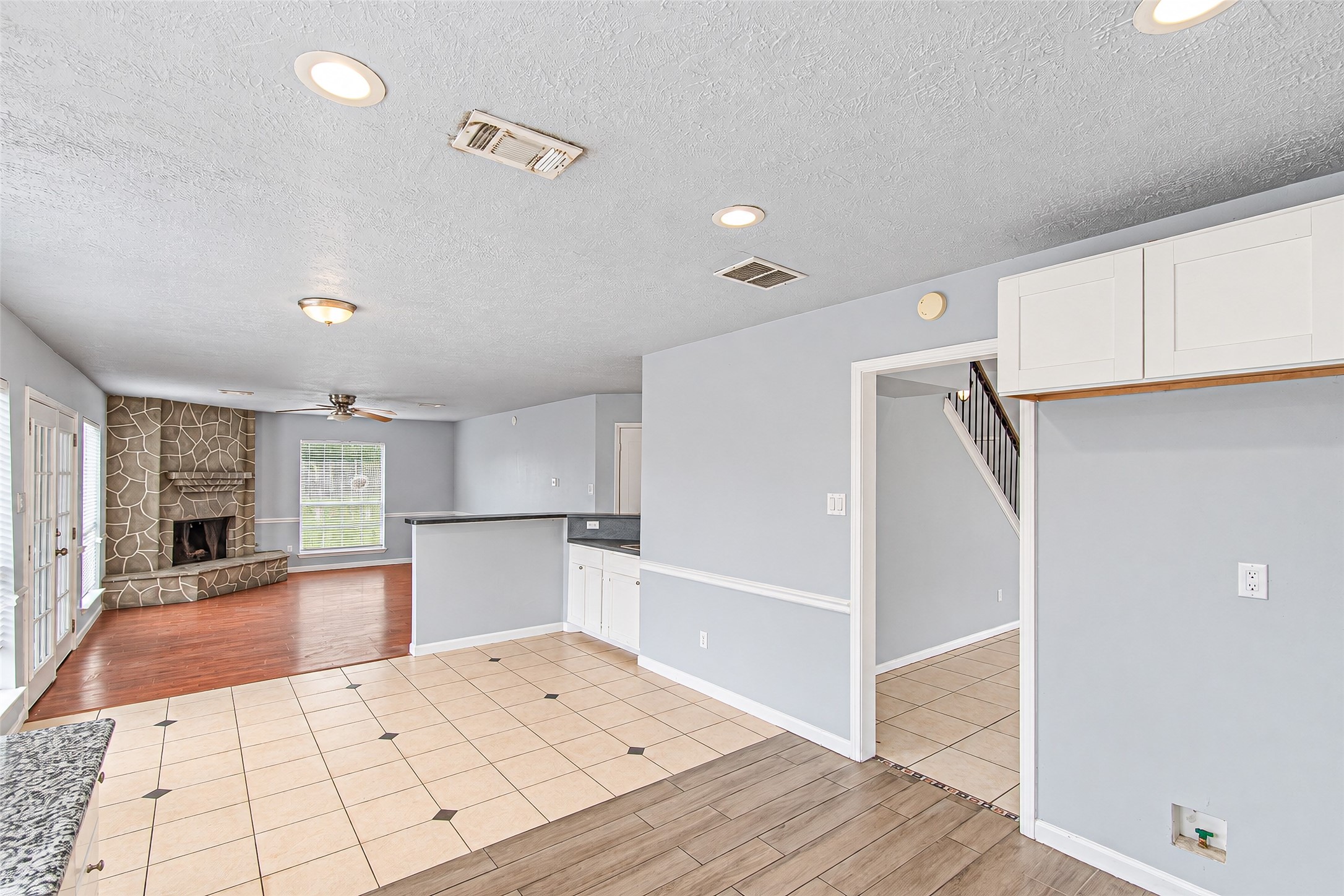 Bright and spacious open-concept living area featuring a cozy stone fireplace, tile and wood flooring, and ample natural light from large windows and French doors.
