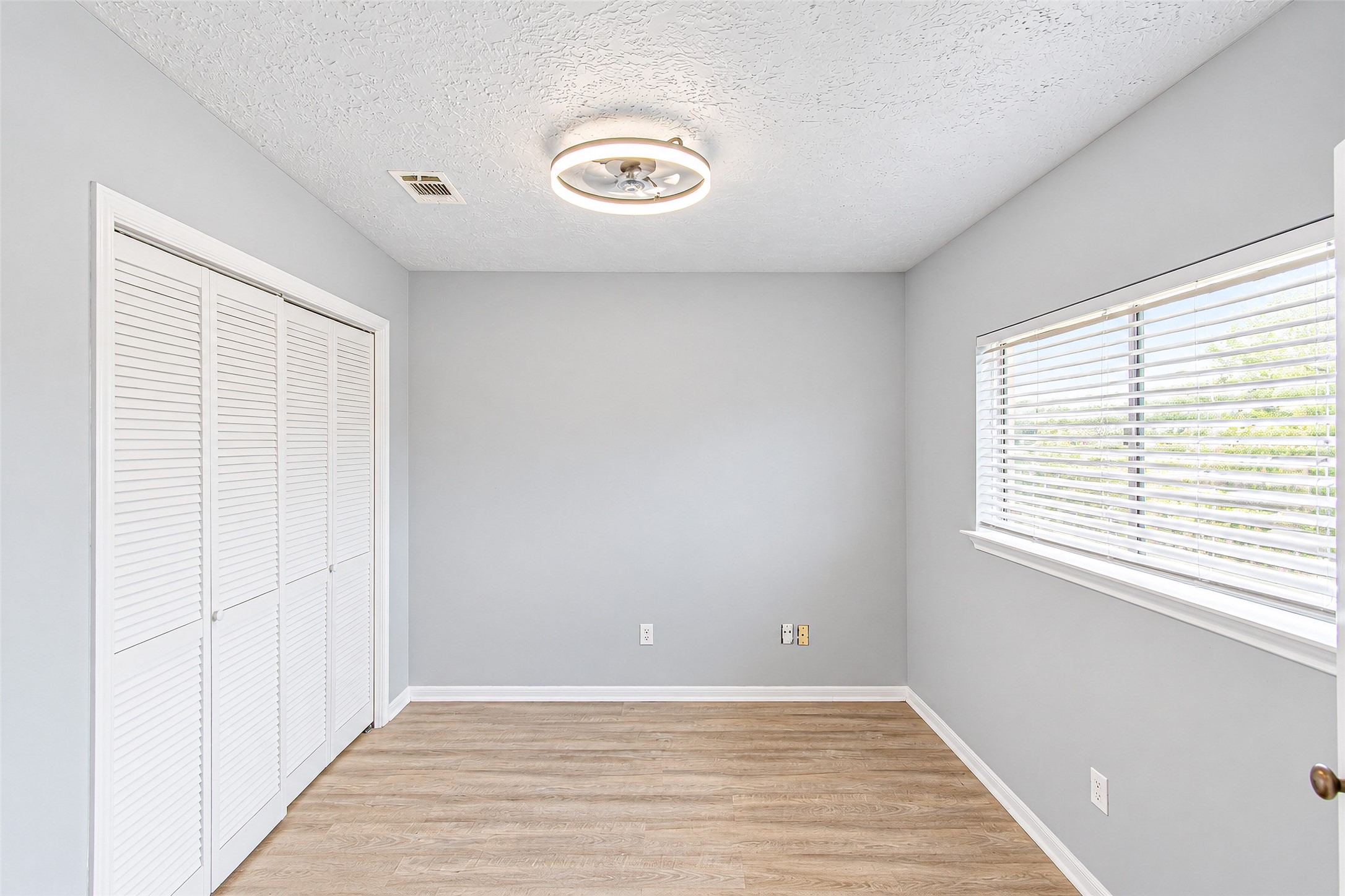 Bright and airy room featuring light hardwood floors, a modern ceiling fixture, and ample closet space with white louvered doors.