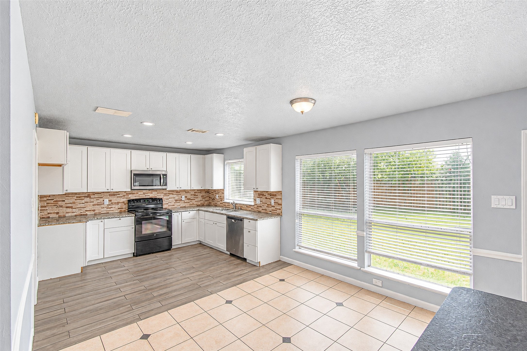 Bright and spacious kitchen with modern white cabinetry, sleek black appliances, and a stylish tile backsplash. Large windows provide abundant natural light and a view of the backyard.