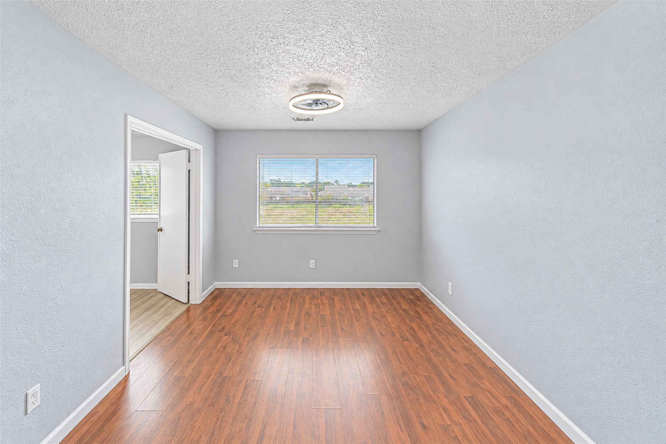 Bright and inviting room featuring sleek hardwood floors, soft gray walls, and a modern light fixture. Abundant natural light flows through the window, highlighting the open layout and creating a warm atmosphere.