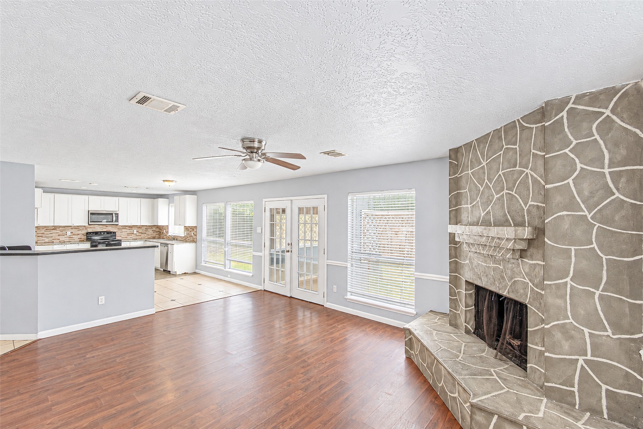 Spacious living area with elegant hardwood floors, a unique stone-patterned fireplace, and abundant natural light from large windows and French doors. The open-concept layout flows seamlessly into a modern kitchen with sleek cabinetry and stainless steel appliances.