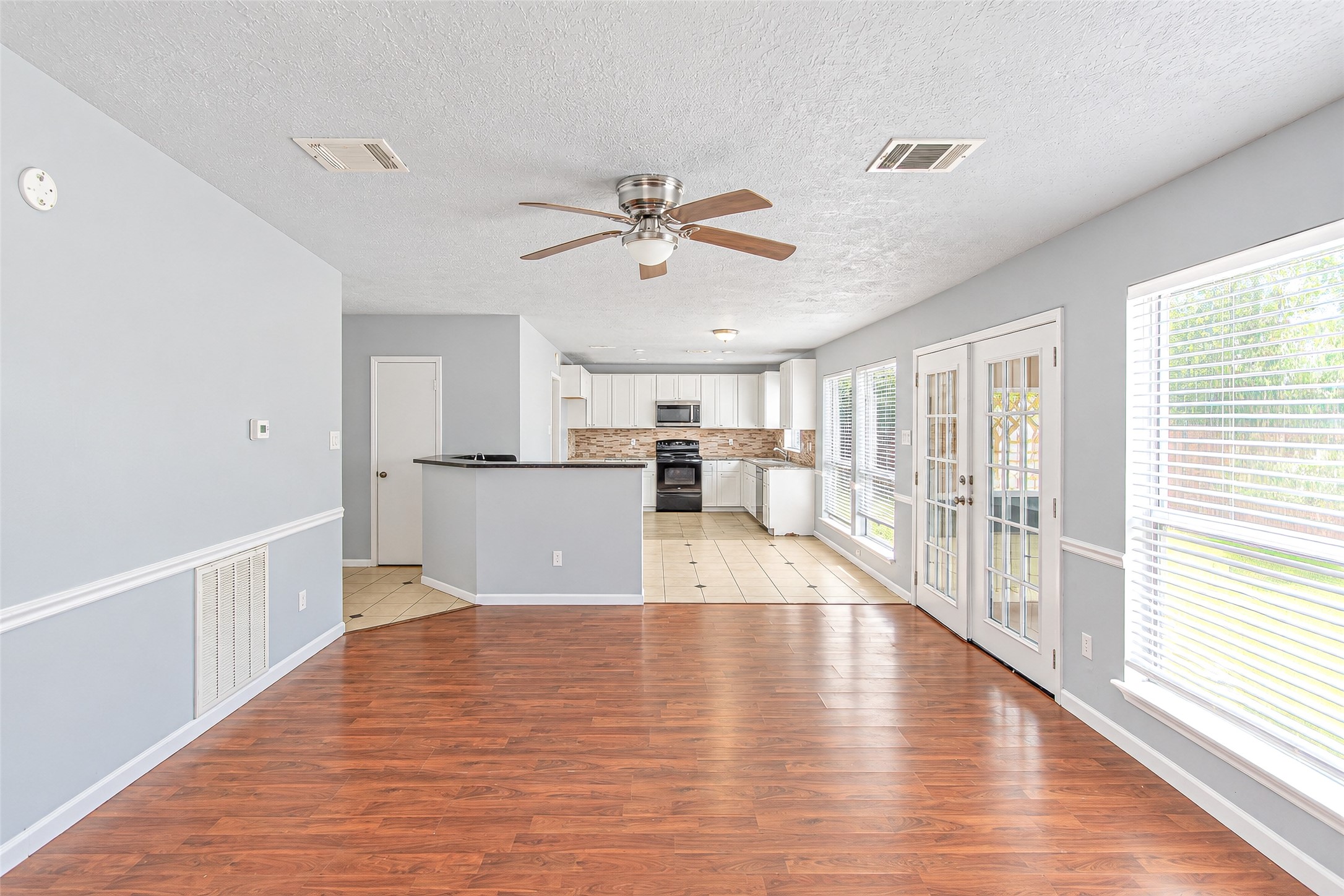 Spacious open-concept living area with elegant wood flooring, abundant natural light through large windows and French doors, and a modern kitchen featuring sleek countertops and stainless steel appliances.