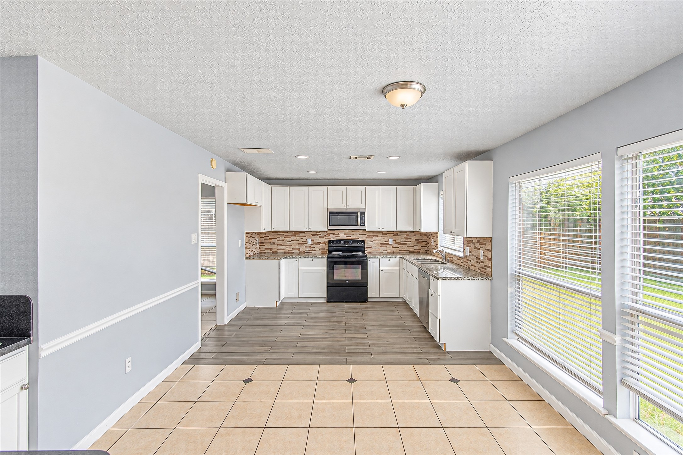Bright and inviting kitchen with sleek white cabinetry, modern black appliances, and a stylish tile backsplash. Large windows provide ample natural light, enhancing the open and spacious feel.