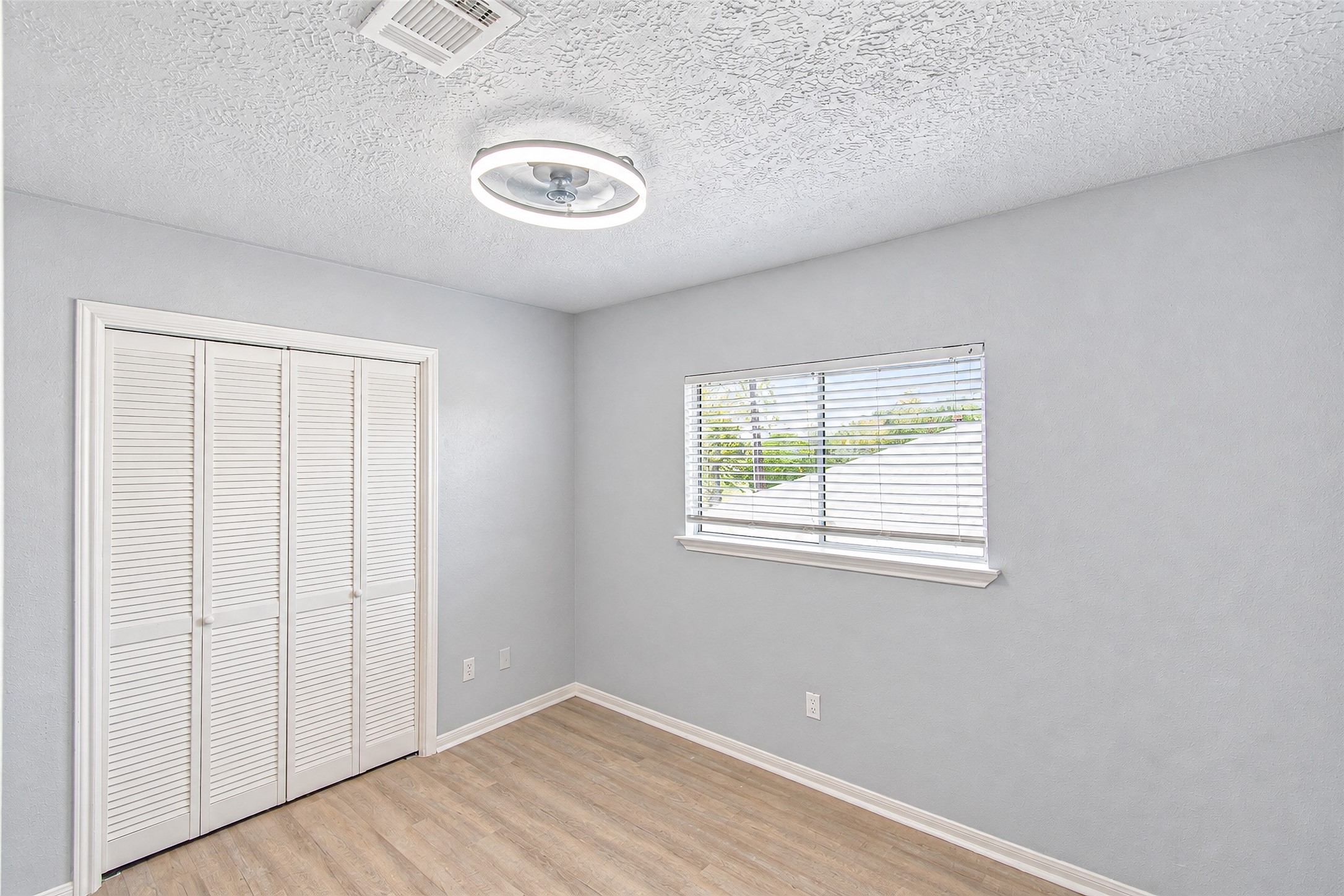 Bright and airy room featuring a modern light fixture, light wood flooring, and a window with blinds, perfect for a cozy retreat.